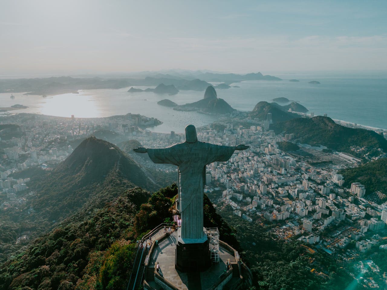 Rear perspective of Christ the Redeemer overlooking Rio de Janeiro skyline with Sugarloaf Mountain and Guanabara Bay at sunset