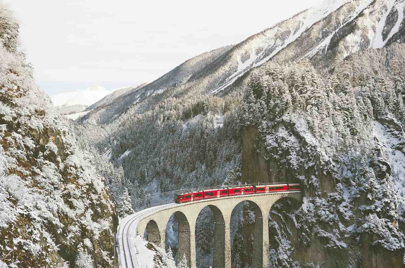 A red train crosses a stone viaduct in a snowy mountainous landscape, surrounded by forest-covered hills.