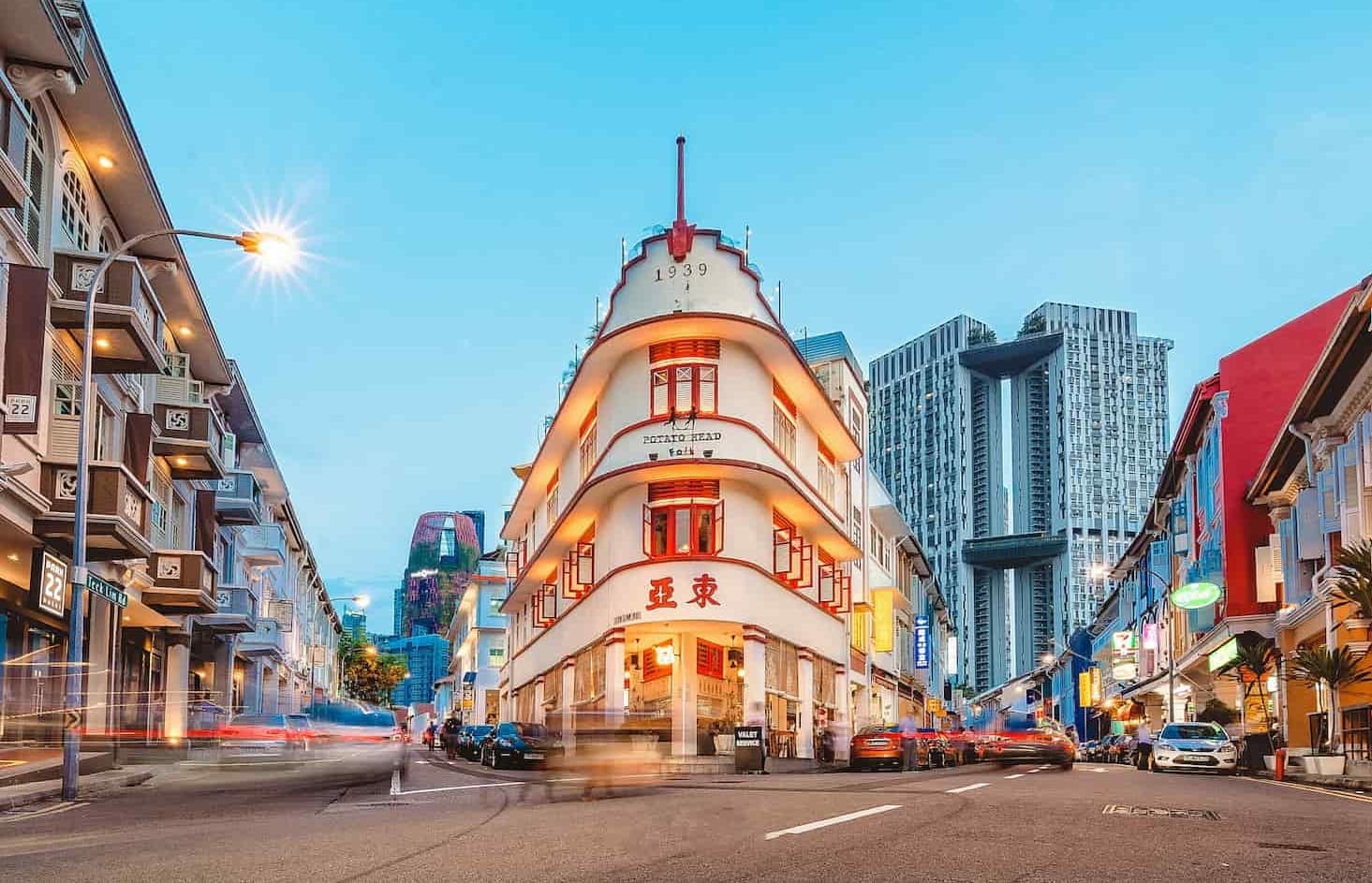 A flatiron-style building with bright lights on a bustling street, surrounded by colorful shophouses and modern high-rise buildings in the background.