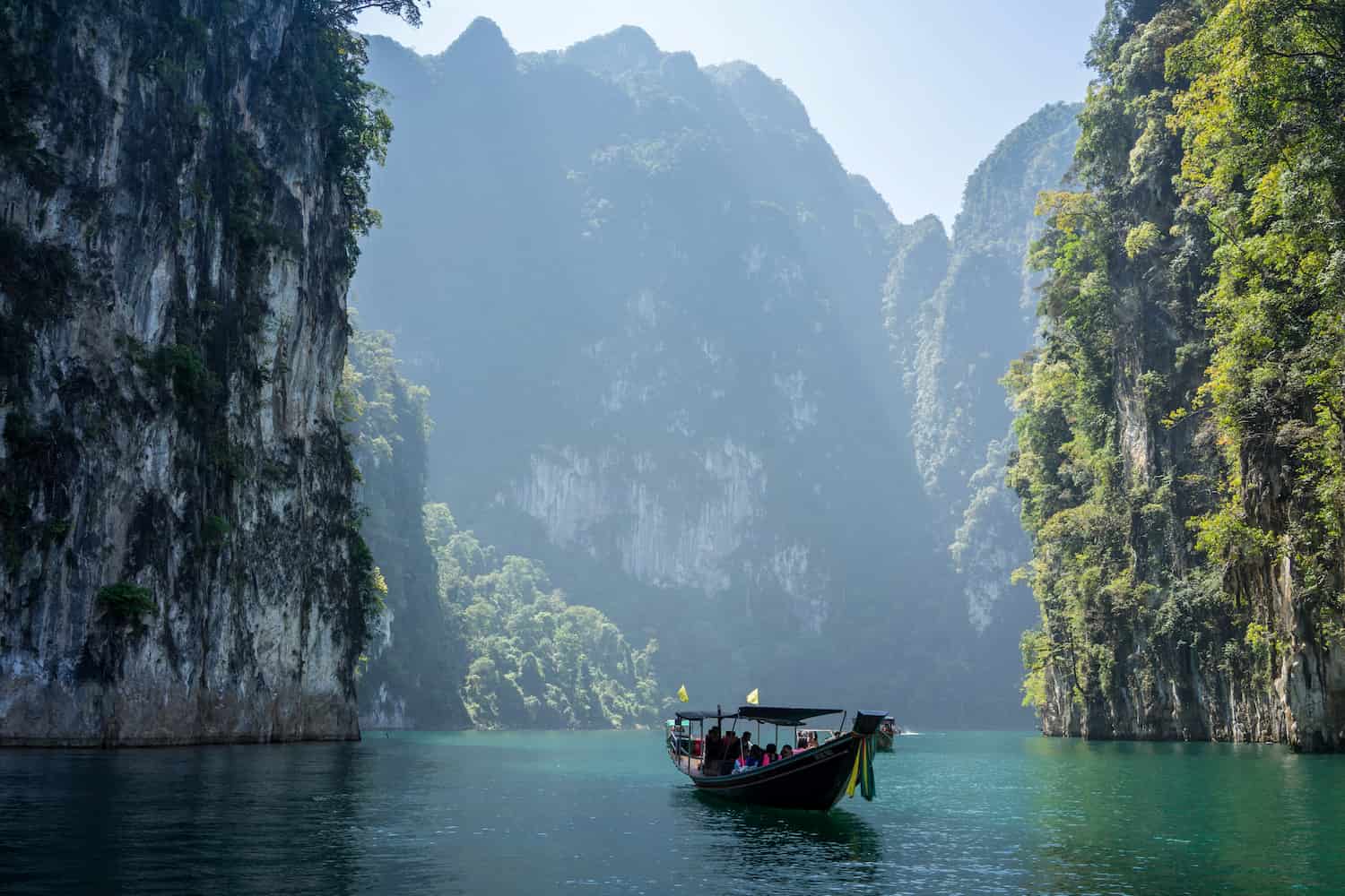 A longtail boat sails through turquoise waters surrounded by towering limestone cliffs with lush greenery under a clear blue sky.