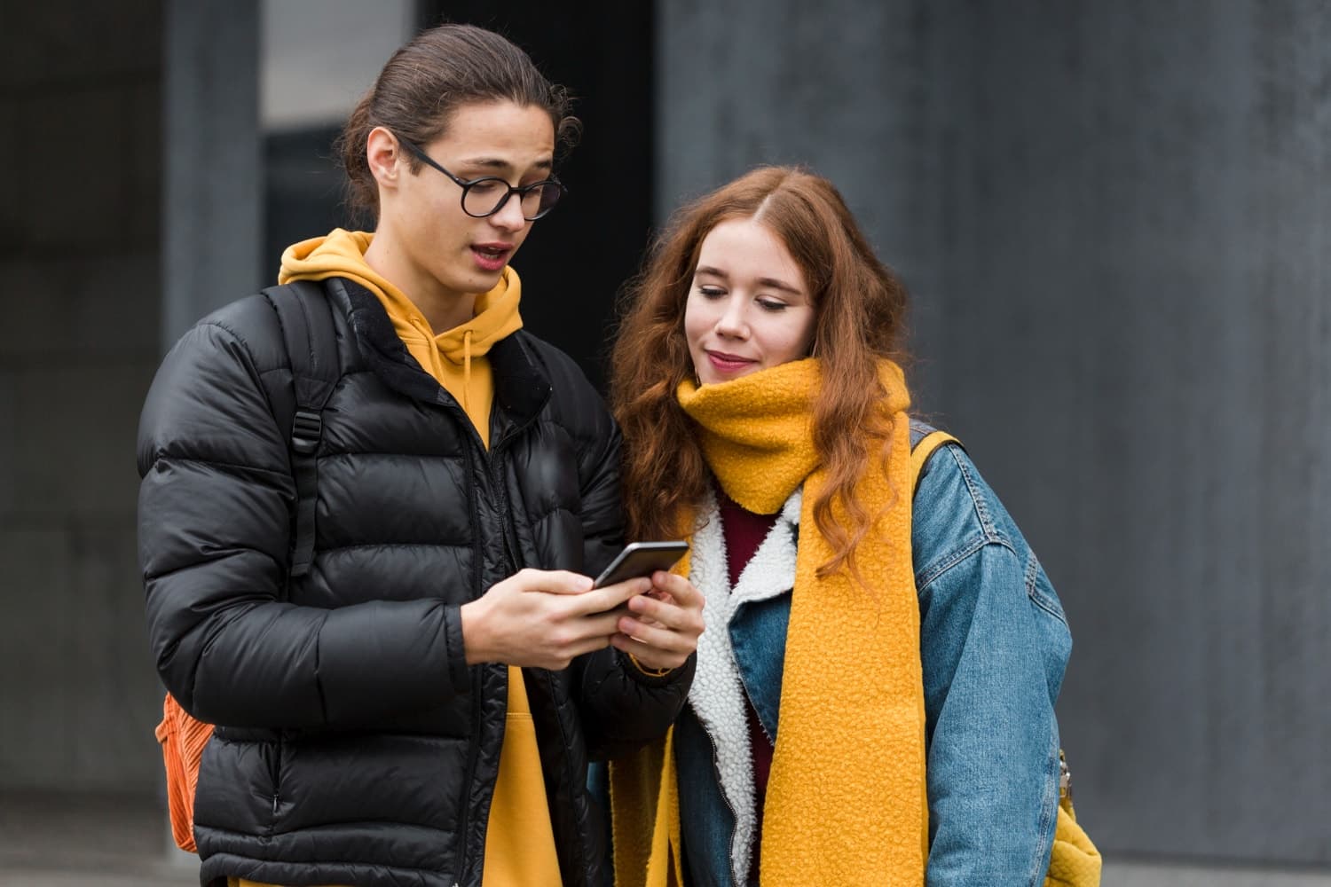 Two people stand together, one holding a smartphone. They wear casual winter clothing, including a yellow scarf and a black jacket.