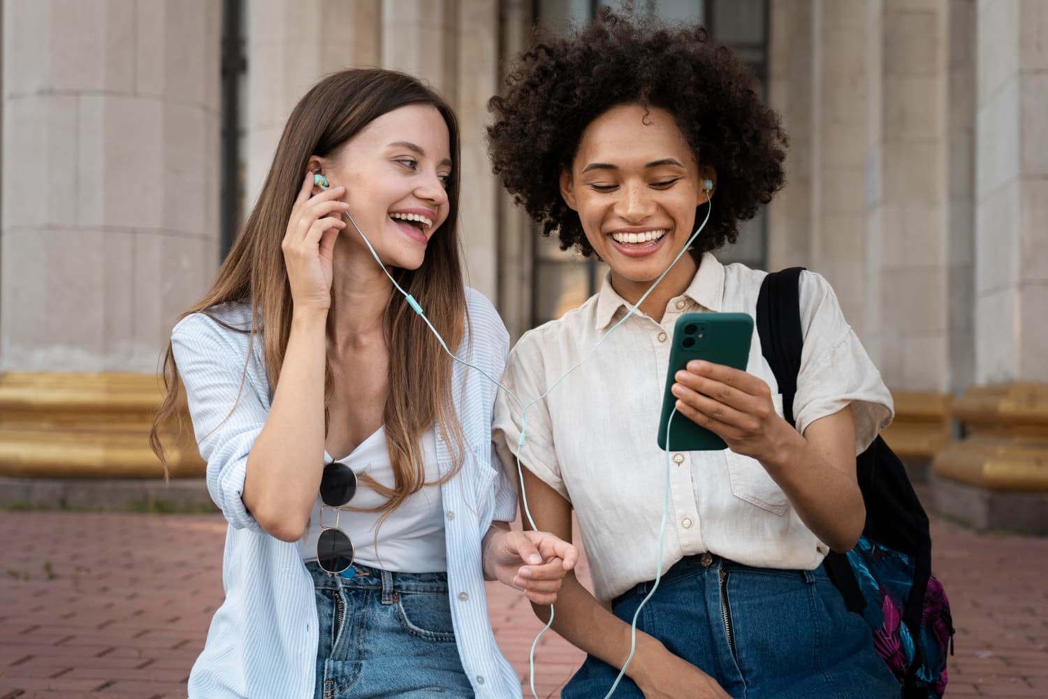 Two women sitting outside, sharing earphones and smiling while looking at a smartphone. One has long hair, the other has curly hair.