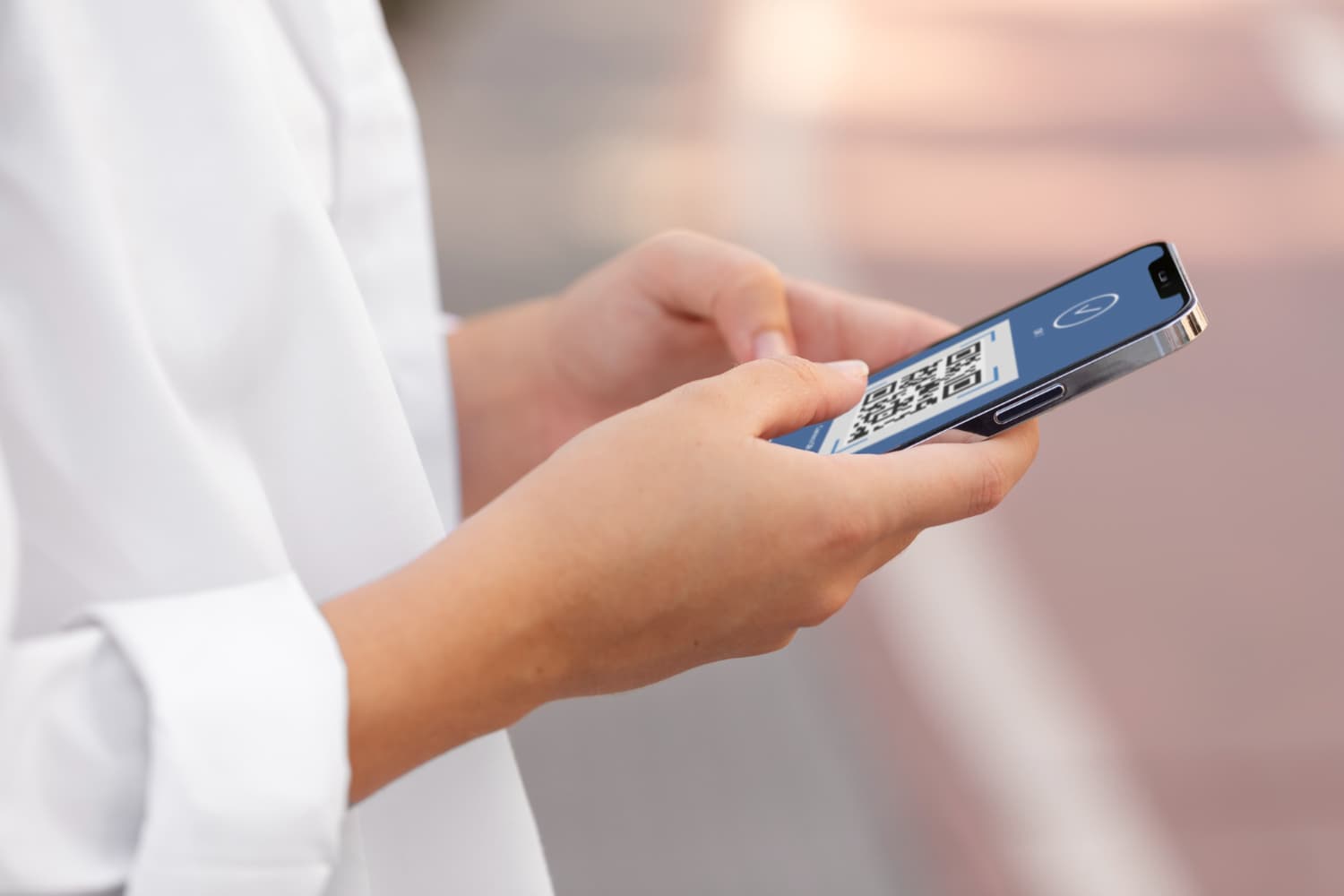 Person in a white shirt holding a smartphone displaying a QR code on the screen, standing outdoors.