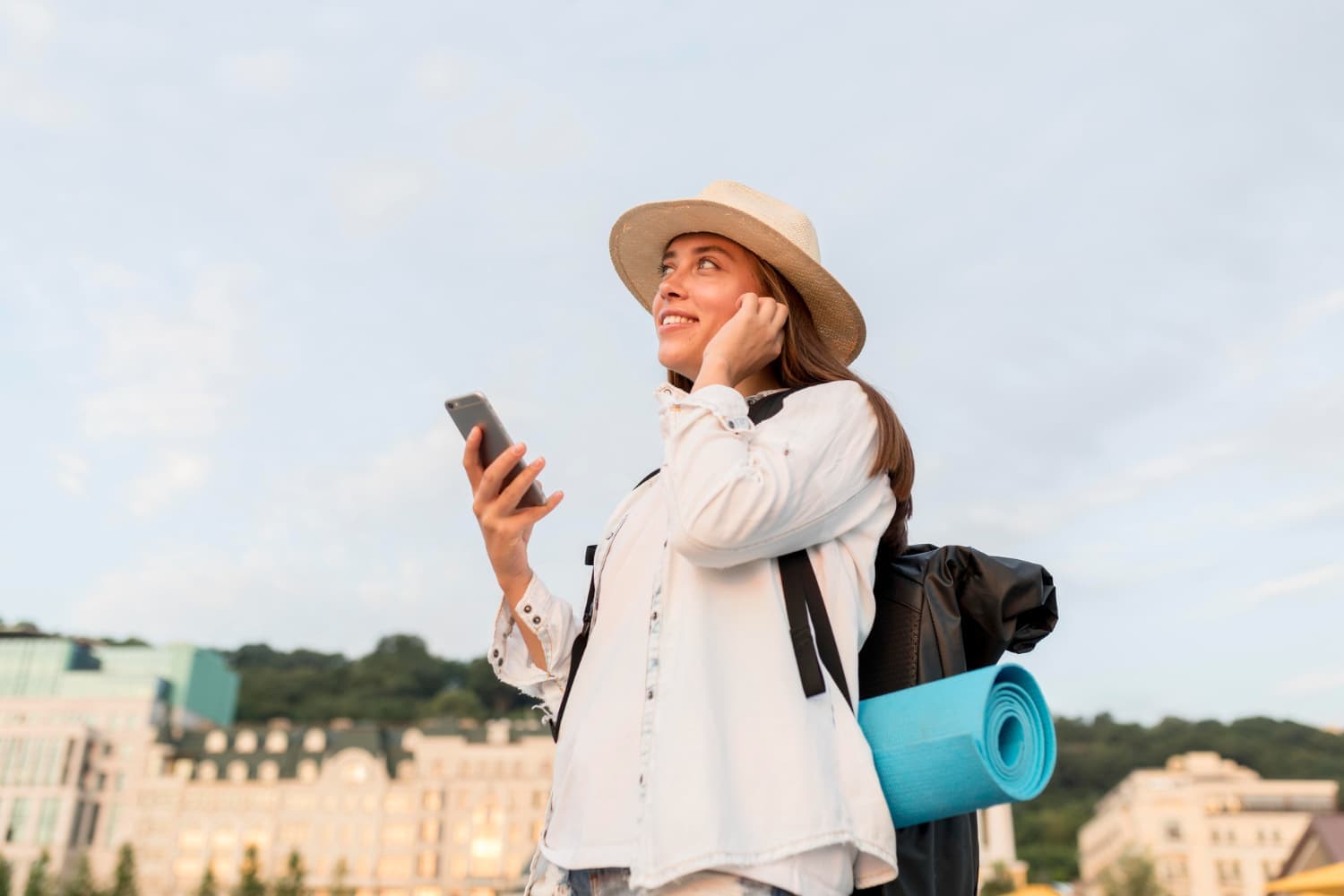 Woman in a hat smiles while holding a smartphone and wearing a backpack with a blue yoga mat, against a cityscape backdrop.