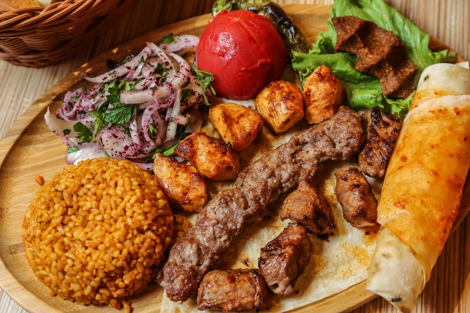 Wooden platter of mixed kebab: lamb kebab skewer, grilled chicken and beef cubes, bulgur, roasted tomato, onion salad, flatbread.