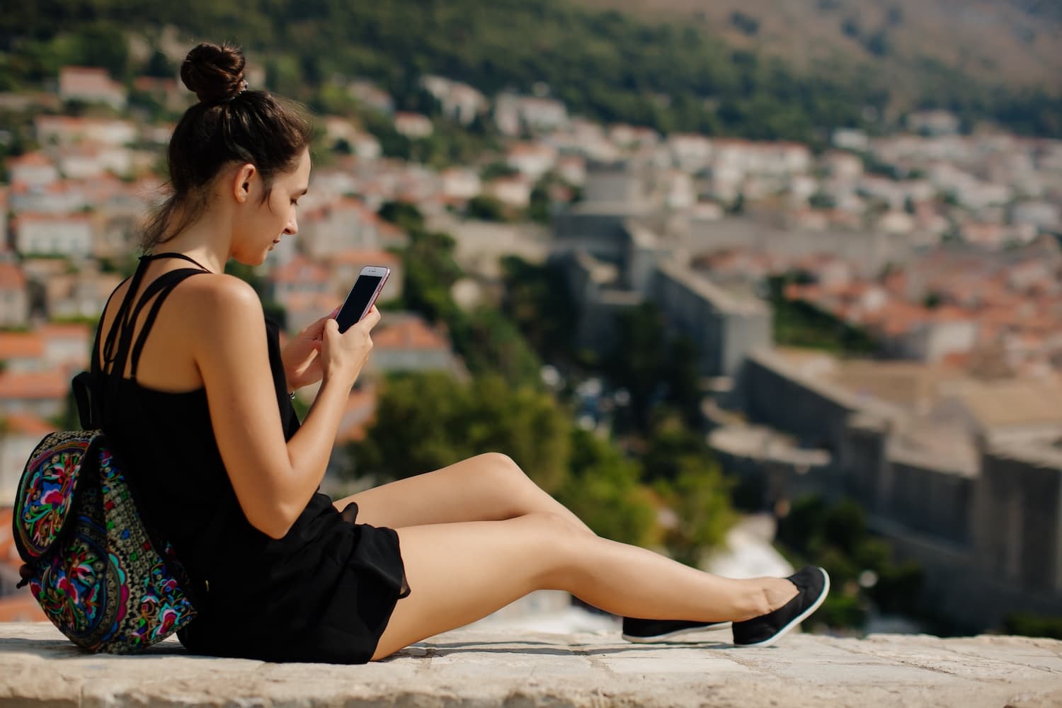 Young woman seated on a stone wall overlooking a red-tiled cityscape, engrossed in her smartphone using free mobile data.