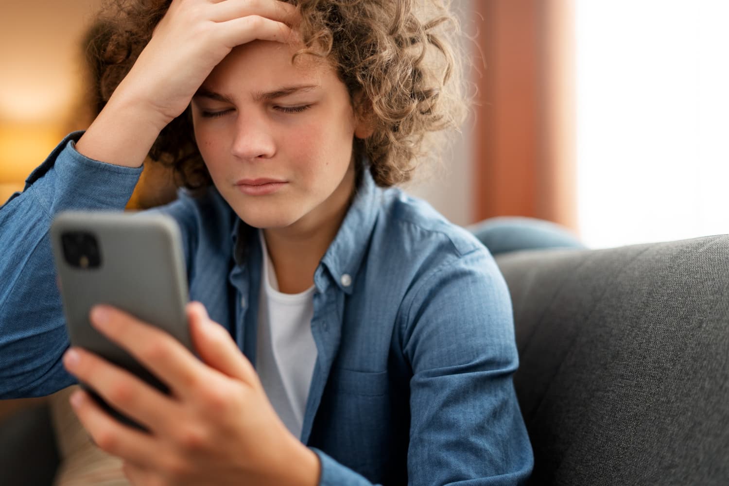 Teen with curly hair sits on a couch, holding a smartphone and looking frustrated, with one hand on their forehead.