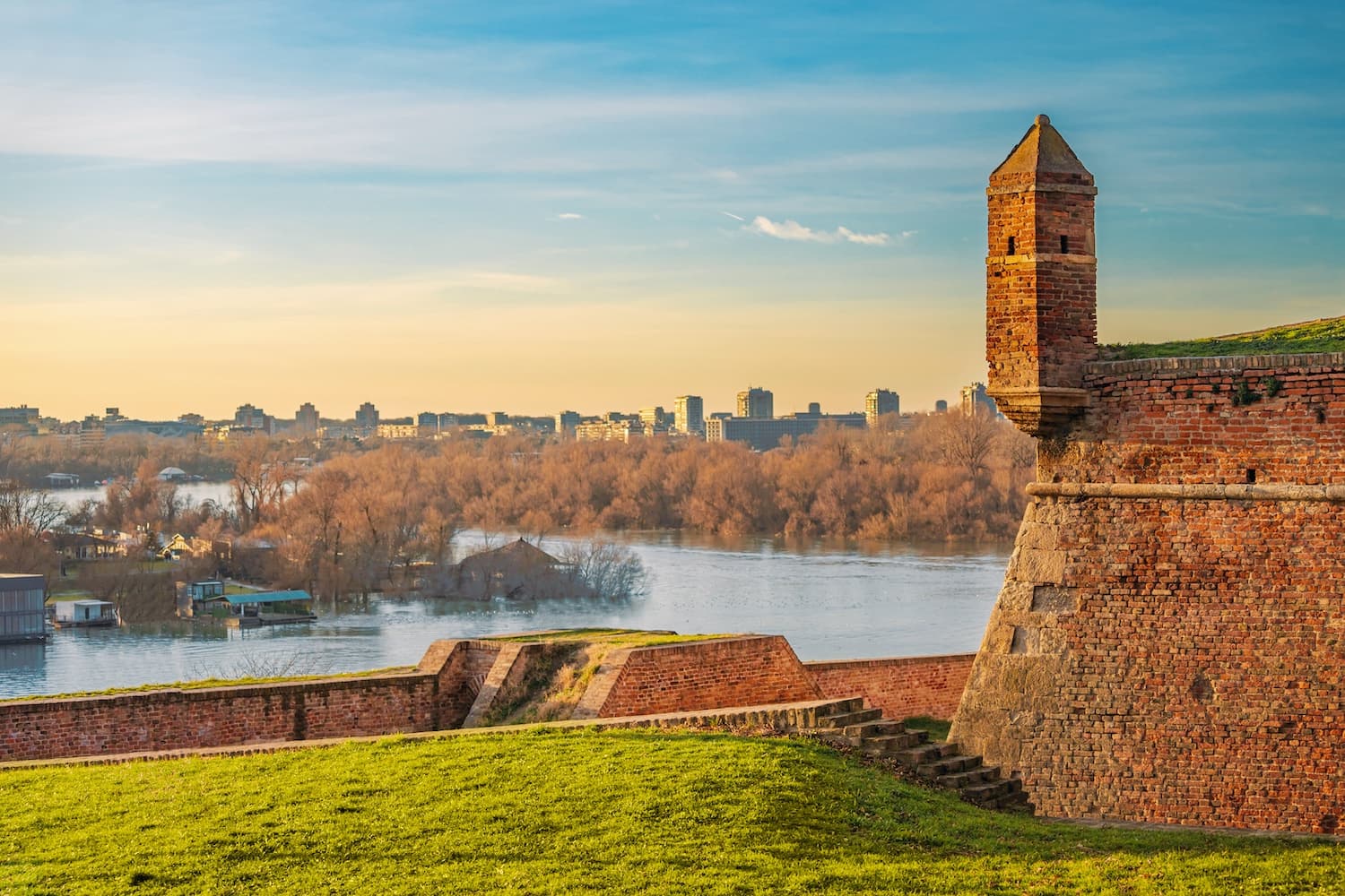 Brick bastion and lookout tower of Belgrade Fortress with grassy embankments, beyond which the Sava River flows past riverside trees and the distant city skyline under a clear sky.