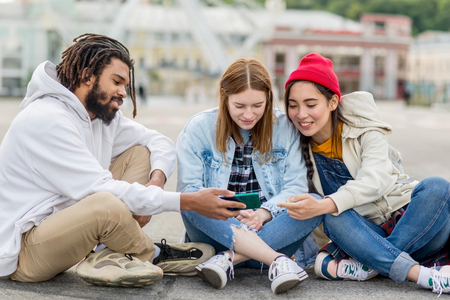 Three friends sitting on the ground, smiling and looking at a smartphone together. They are casually dressed and appear to be enjoying themselves.
