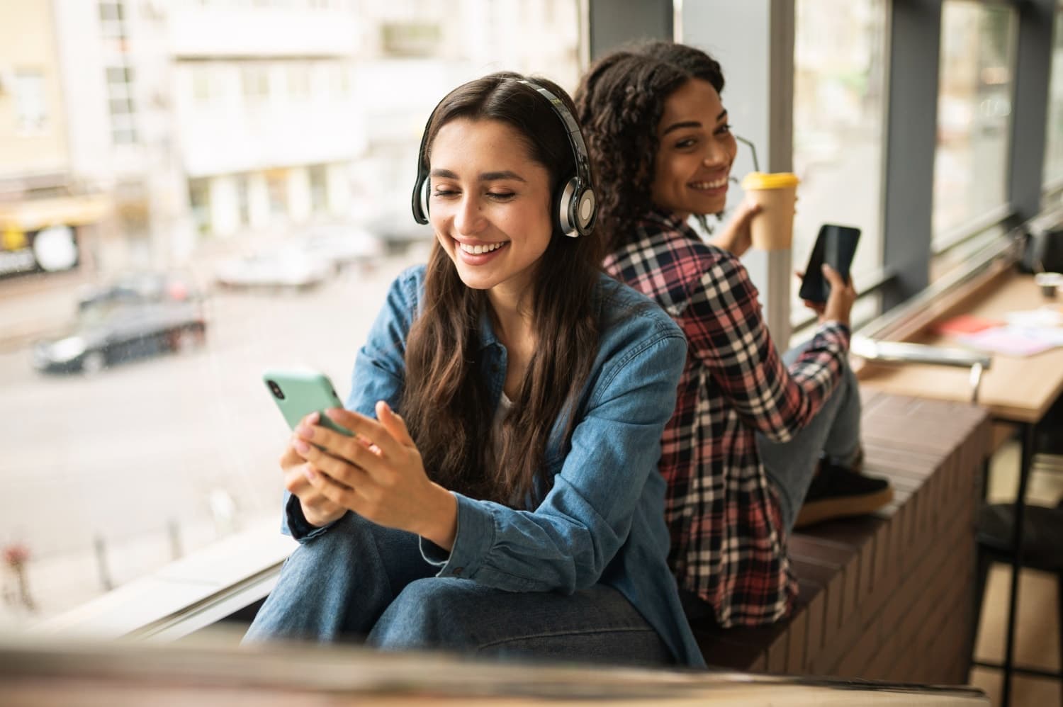 Two students checking their phones together by a cafe window