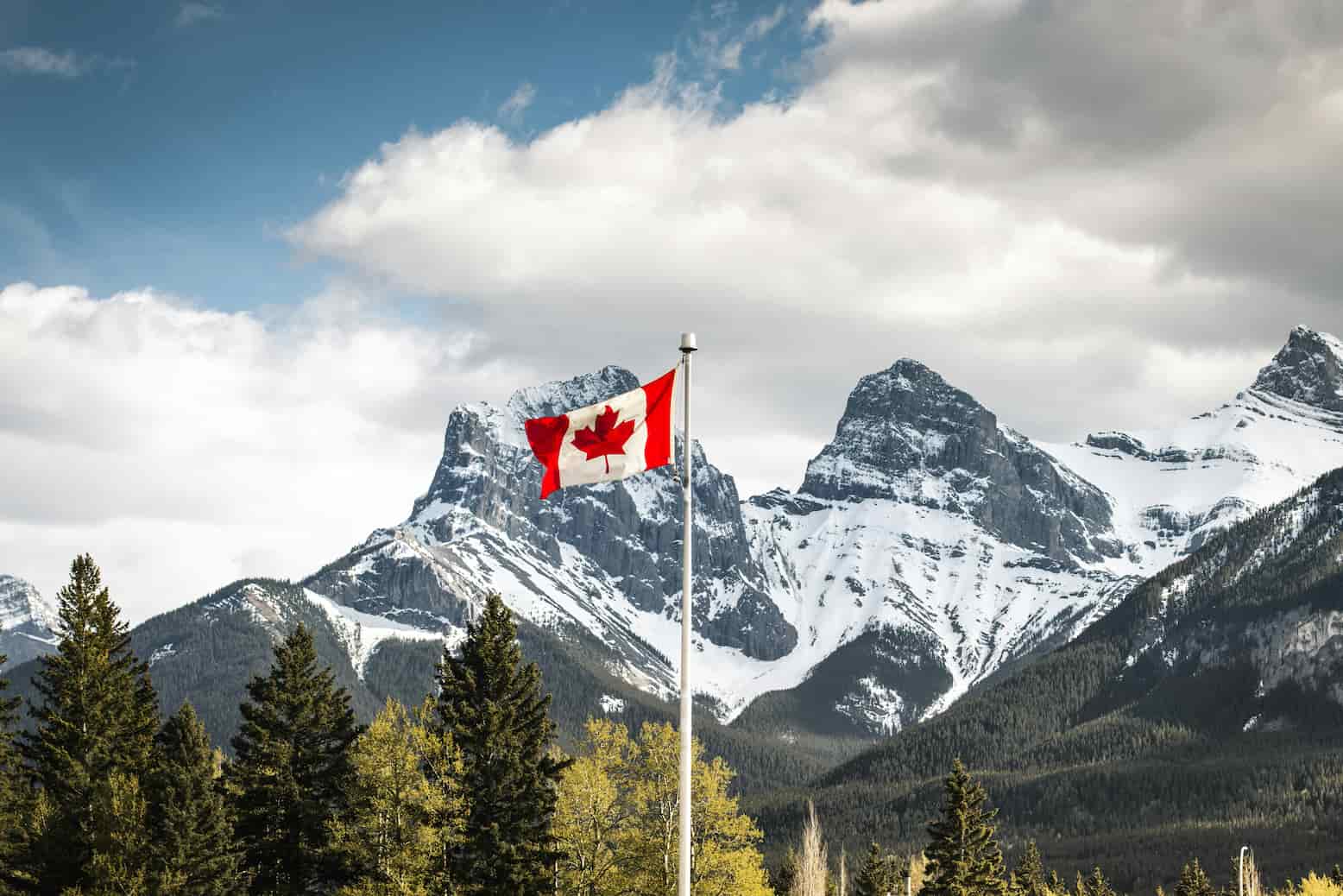 Canadian flag waving in front of snow-capped mountains and forest under a partly cloudy sky.