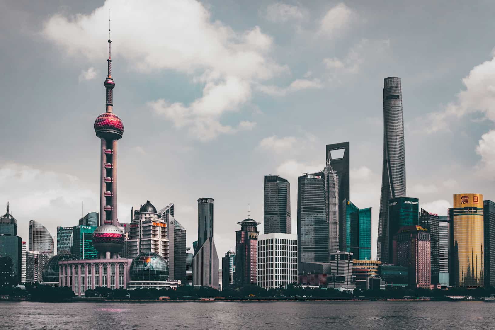 Shanghai skyline featuring the Oriental Pearl Tower and modern skyscrapers under a cloudy sky, viewed from across the river.
