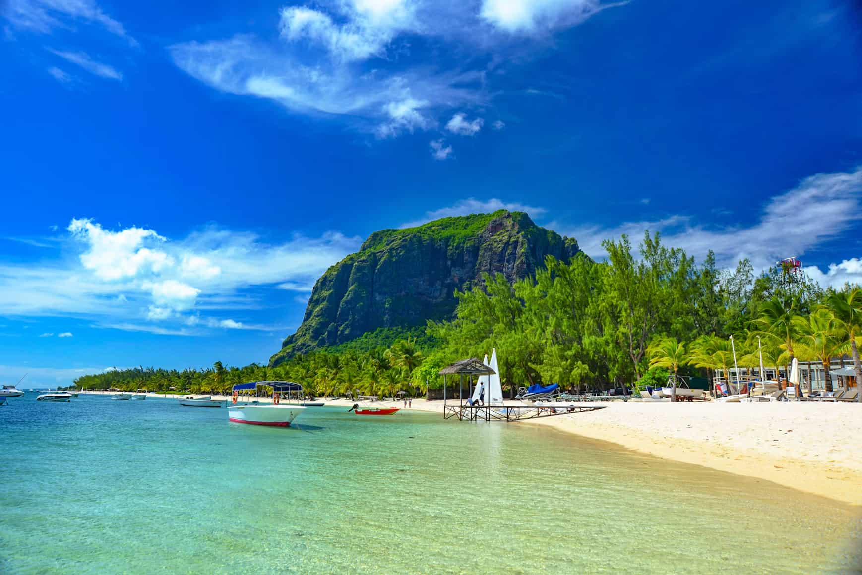 Clear turquoise water, sandy beach, lush green trees, boats, and a towering mountain under a vibrant blue sky.