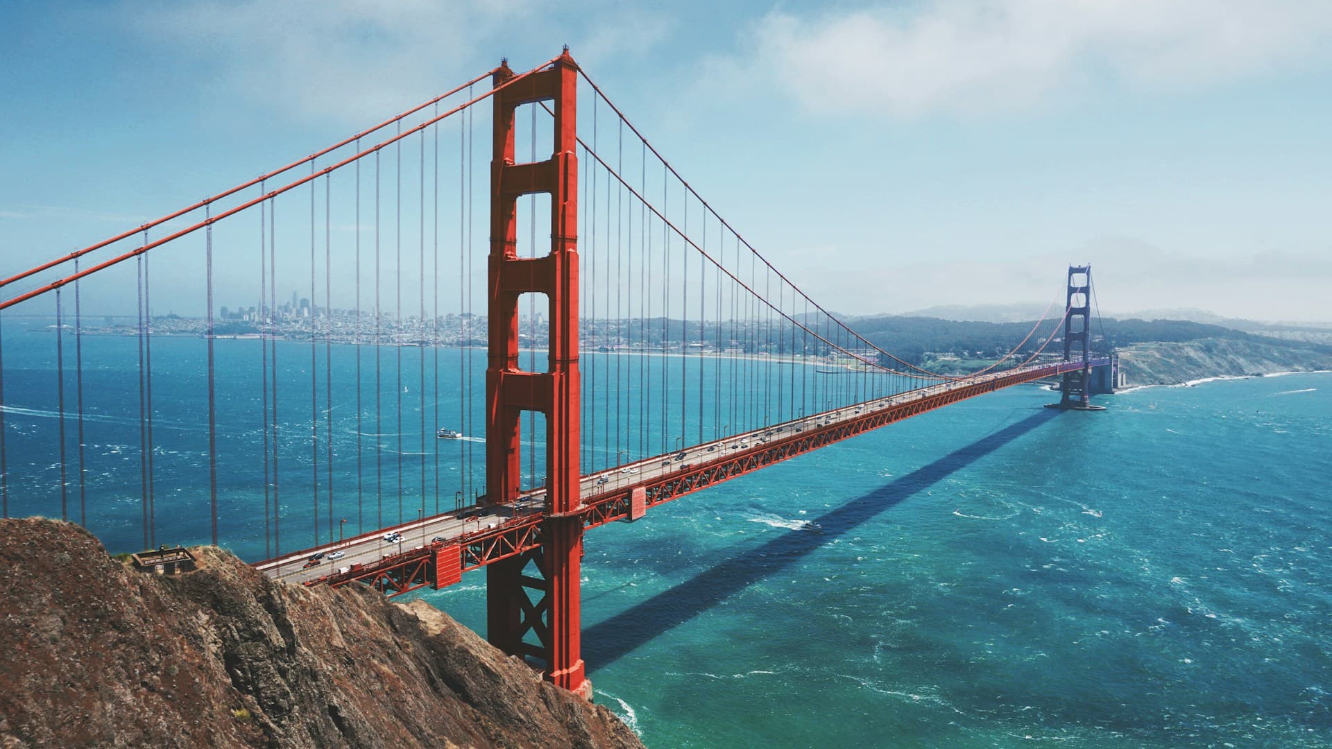 Aerial view of the Golden Gate Bridge spanning blue waters, with a clear sky and distant cityscape in the background.