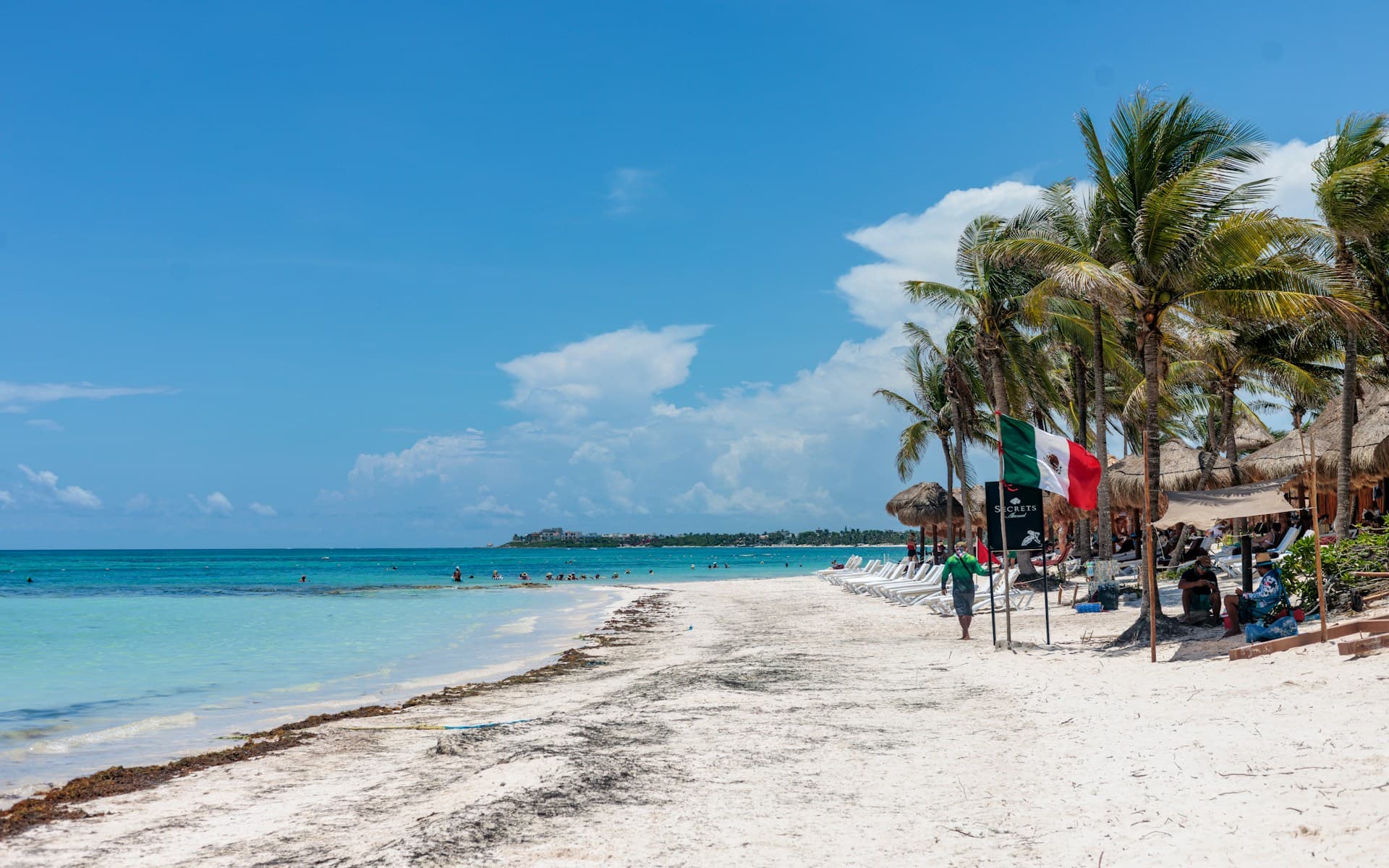 Sunny beach with palm trees, turquoise sea, and people relaxing. Mexican flag on display. Clear blue sky and fluffy clouds in the background.