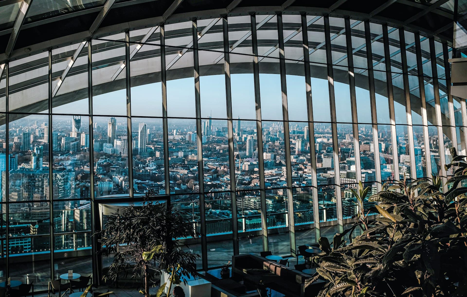 Interior view of a city skyline through curved floor-to-ceiling glass windows, with indoor plants and seating in the foreground.