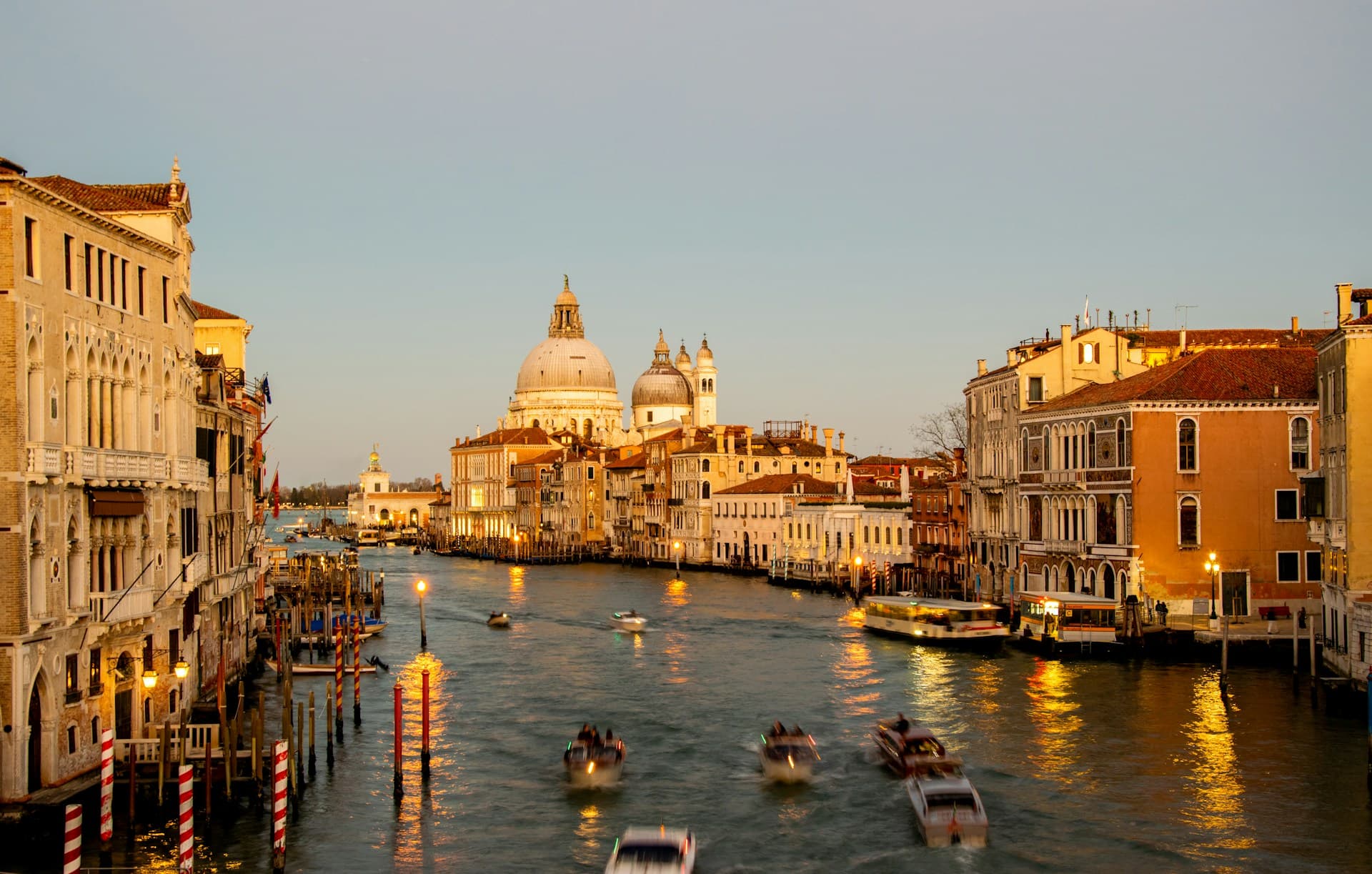 Venice's Grand Canal at sunset, with boats gliding on the water, historic buildings lining the canal, and a prominent dome in the background.