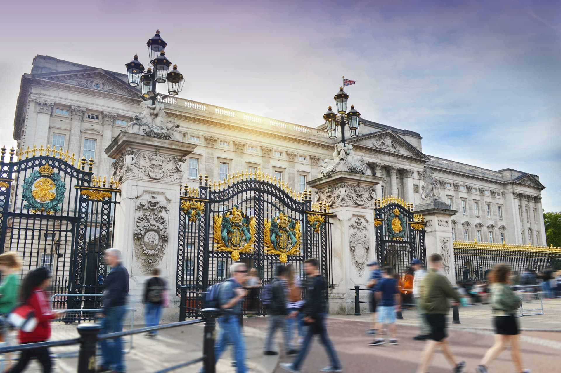 Buckingham Palace facade behind ornate gold-accented iron gates, with blurred pedestrians walking past under a soft sky.