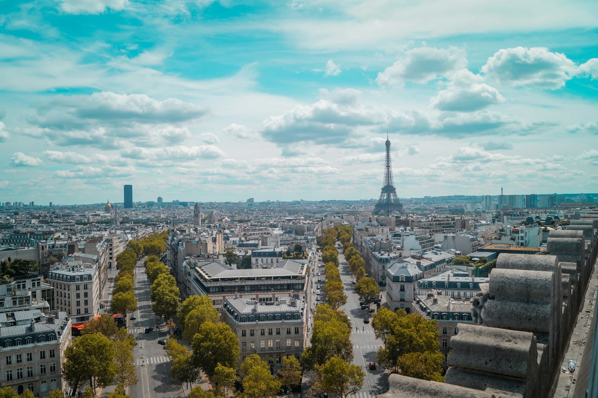 Aerial view of Paris with the Eiffel Tower in the distance, surrounded by city buildings and tree-lined streets under a partly cloudy sky.