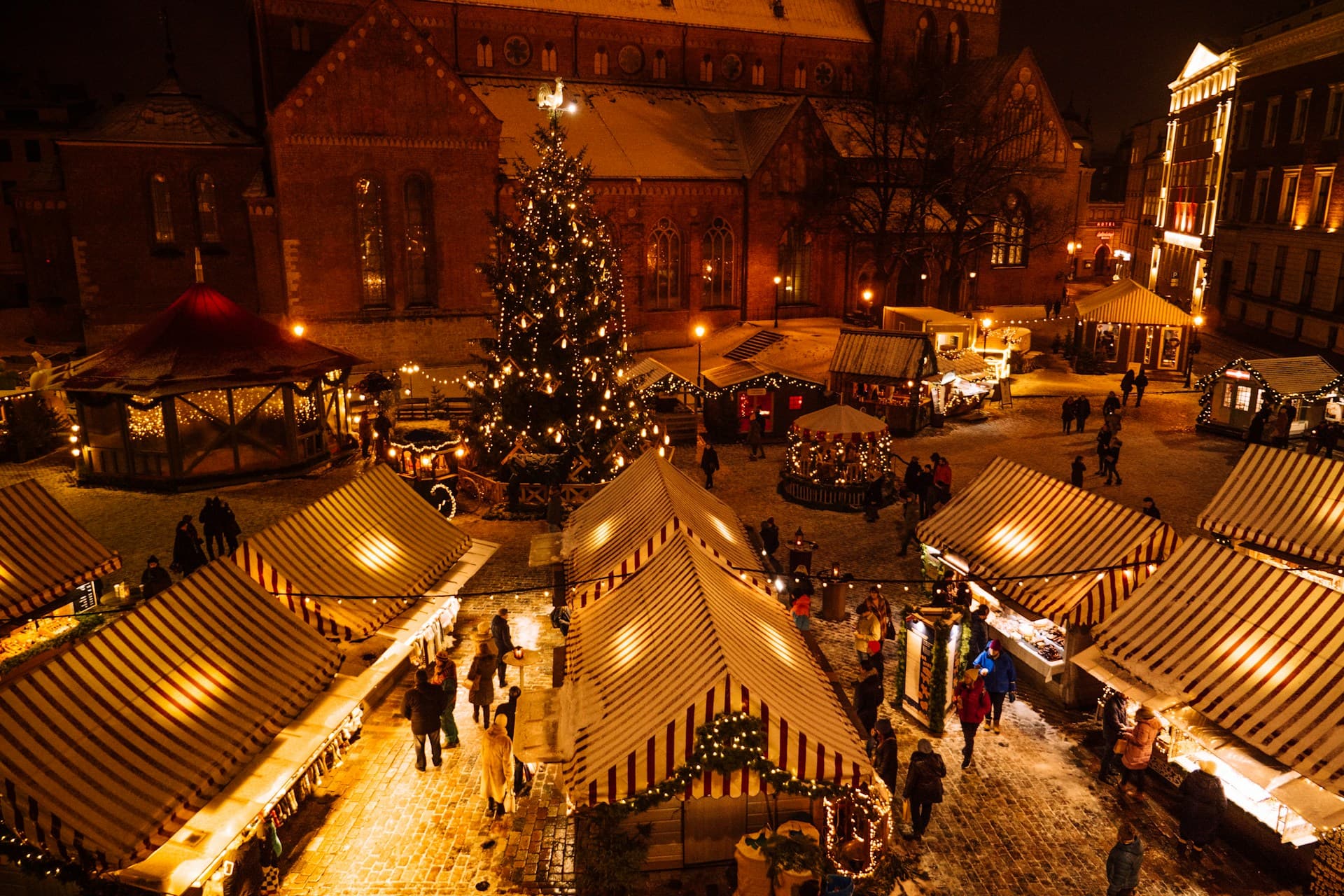 Aerial view of a festive Christmas market at night, with illuminated stalls, a large decorated tree, and people walking on snow-covered paths.