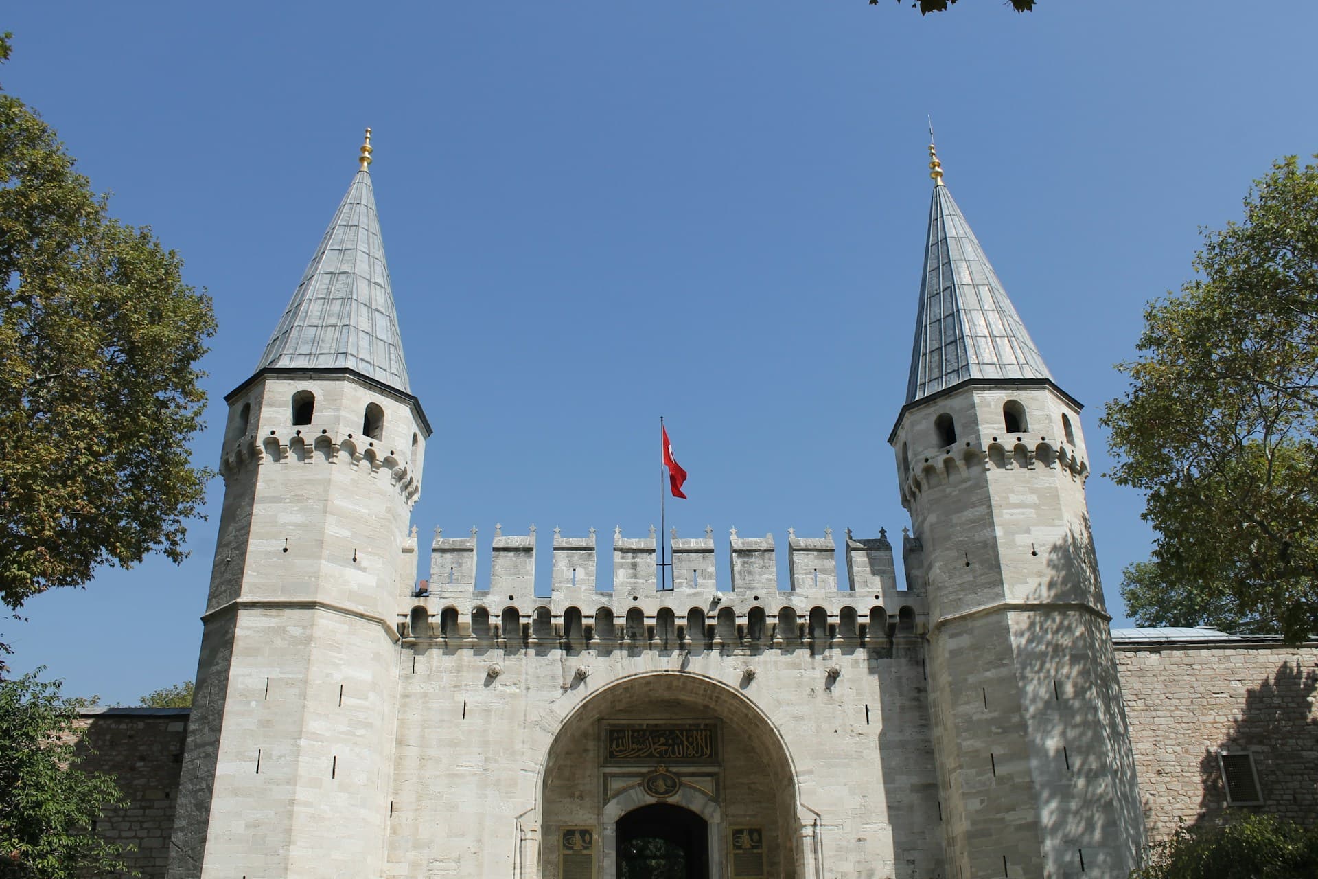 Stone imperial gate with twin conical towers, central arched entrance and Turkish flag overhead against clear blue sky, trees at sides.
