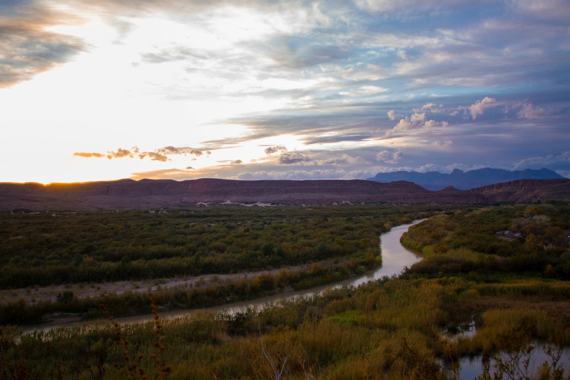 Sunset over a winding river with lush greenery, distant mountains, and a dramatic sky with clouds.