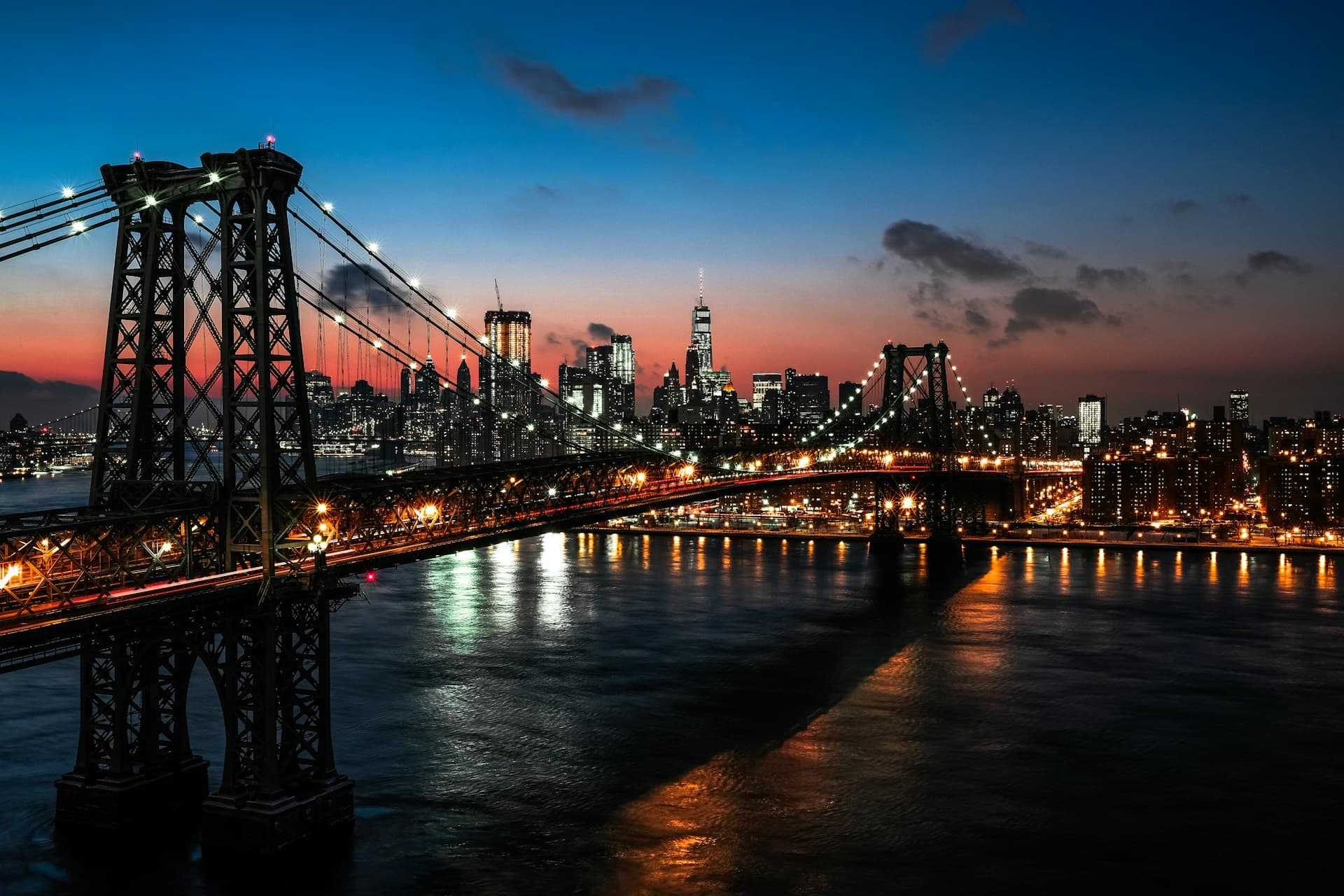 Illuminated city skyline and bridge at dusk, with reflections on the water and a colorful sky transitioning from blue to orange.