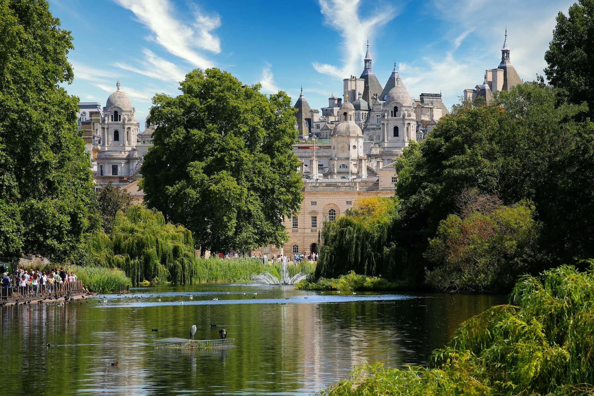 Pond with fountain and ducks in a leafy park, framed by trees and ornate historic stone buildings under a bright blue sky.