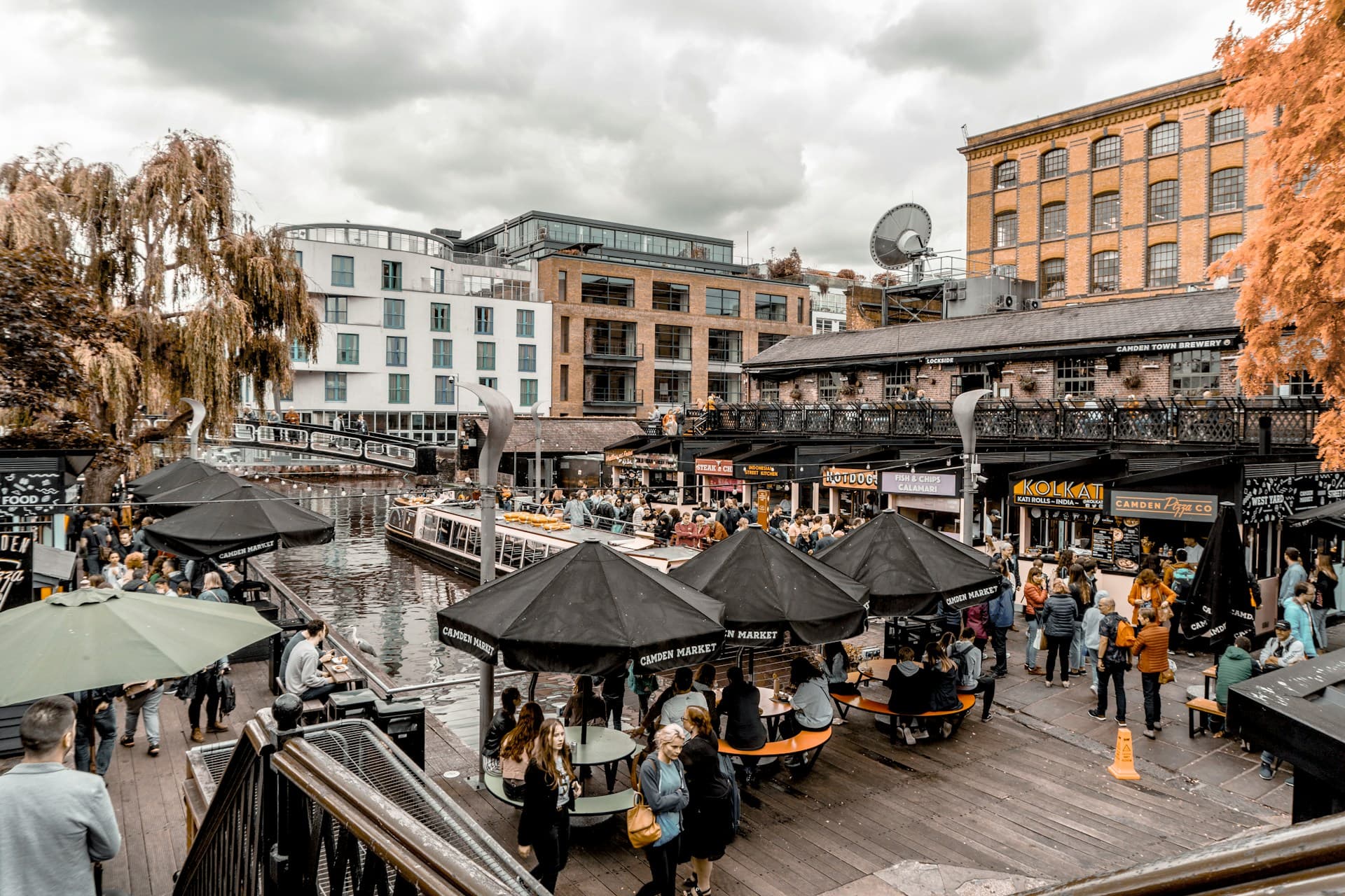 Busy canal-side market with outdoor seating under black umbrellas, boats on the water and brick buildings under a cloudy sky.