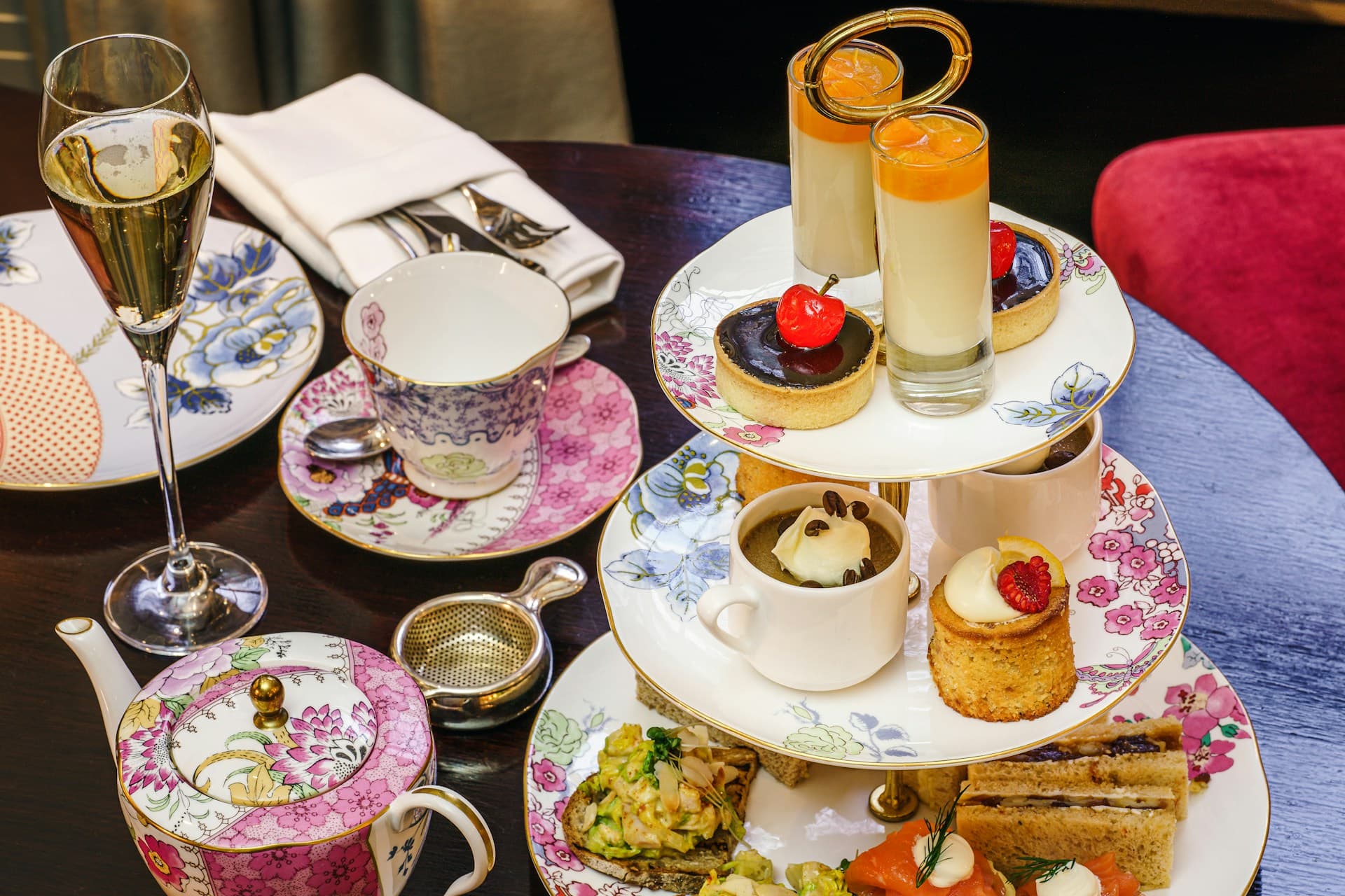 Three-tier afternoon tea with sandwiches, scones and desserts on floral china, teapot and champagne flute on table.