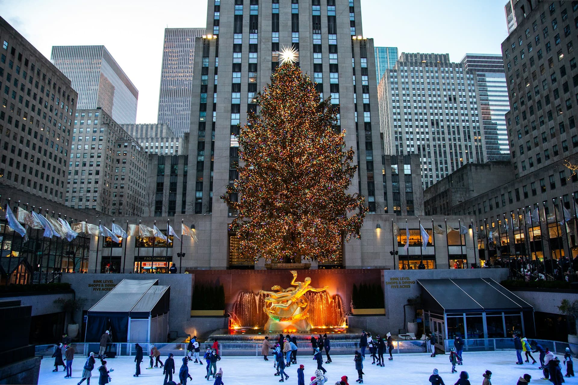 Skaters glide on an ice rink at Rockefeller Center, NYC, beneath a large, illuminated Christmas tree and surrounding tall buildings.