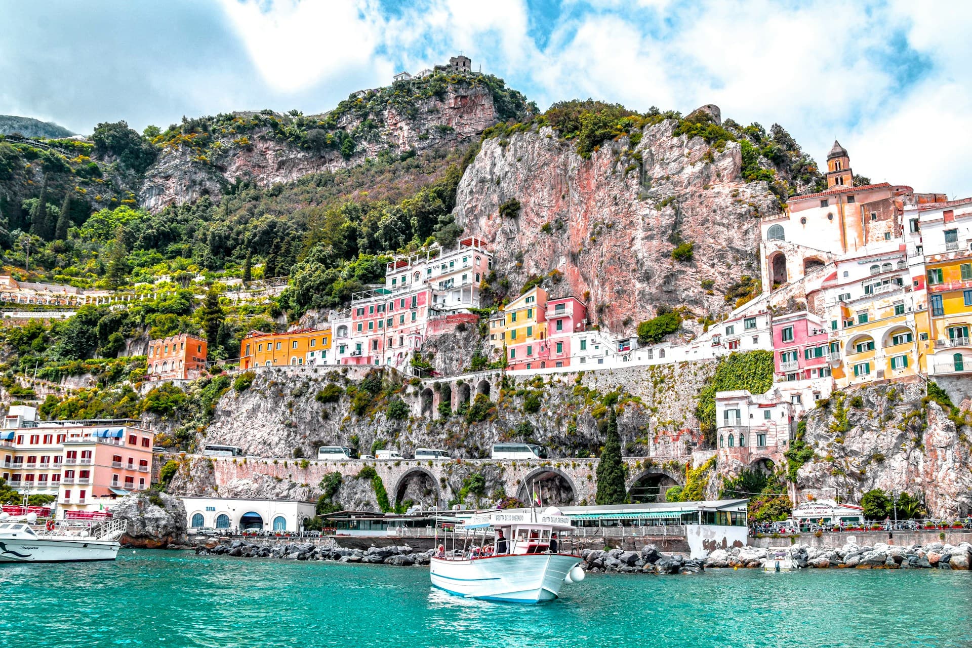 Colorful houses on a rocky hillside overlook a turquoise sea with boats, under a partly cloudy sky.