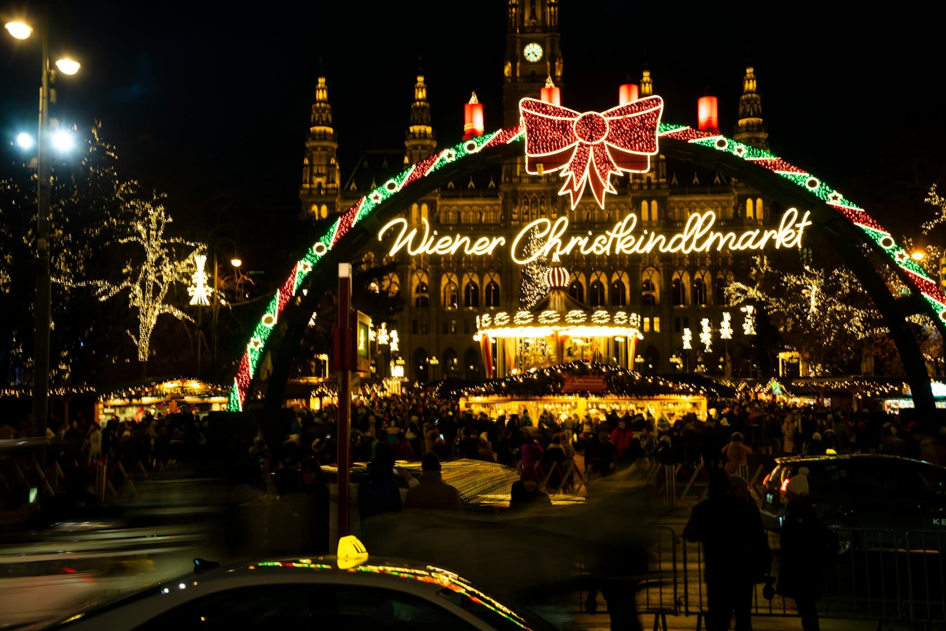 Festive Christmas market at night, with illuminated archway reading "Wiener Christkindlmarkt" and a large building in the background.