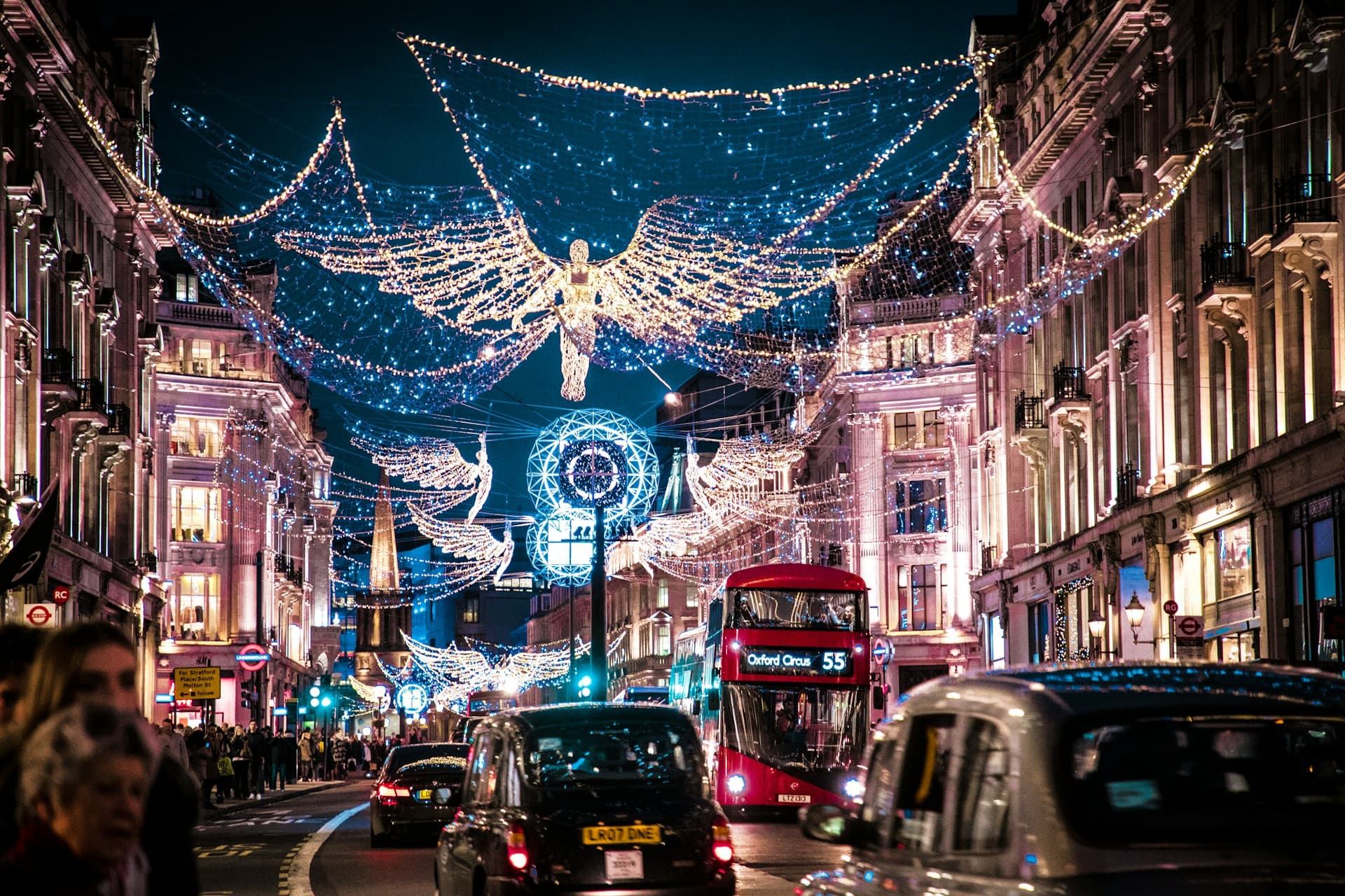 A bustling city street at night, adorned with festive angel-shaped lights. Red buses and taxis move among illuminated buildings.