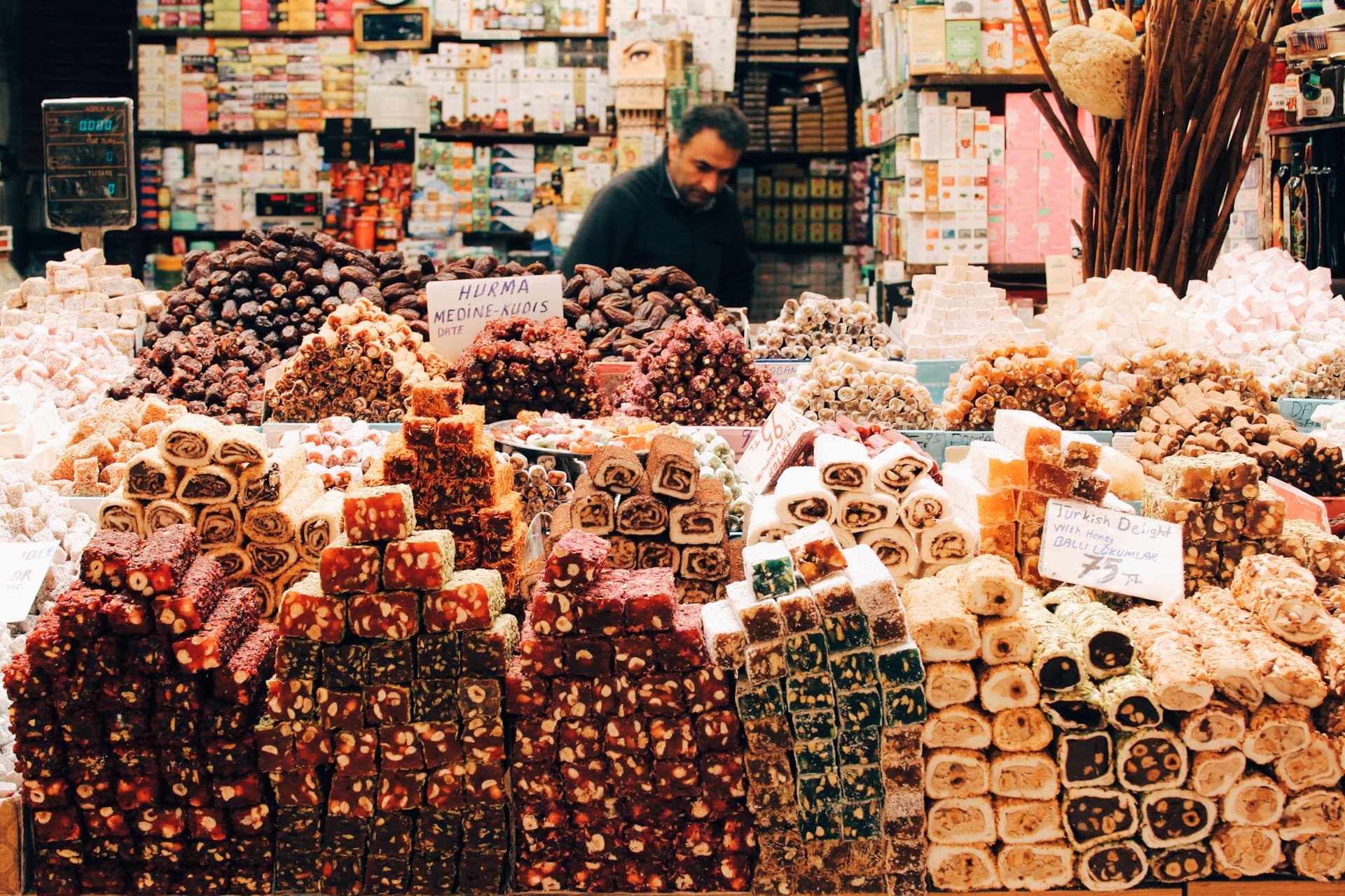 Colorful piles of Turkish delight and dried fruits stacked in pyramids at a bustling market stall, vendor arranging goods behind.