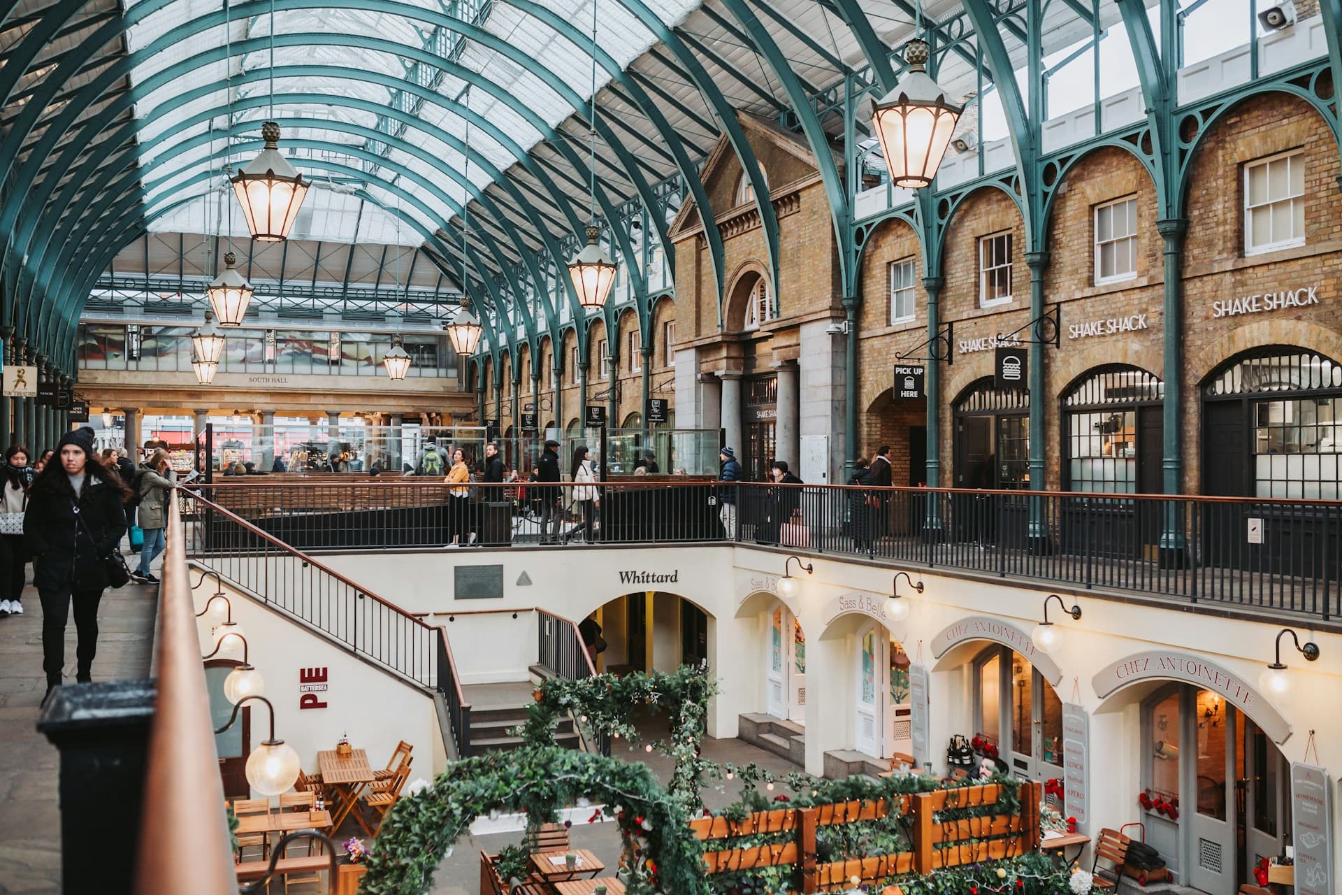 Victorian-style indoor market with arched glass roof, two levels of shops and cafes, hanging lanterns and shoppers walking and browsing.