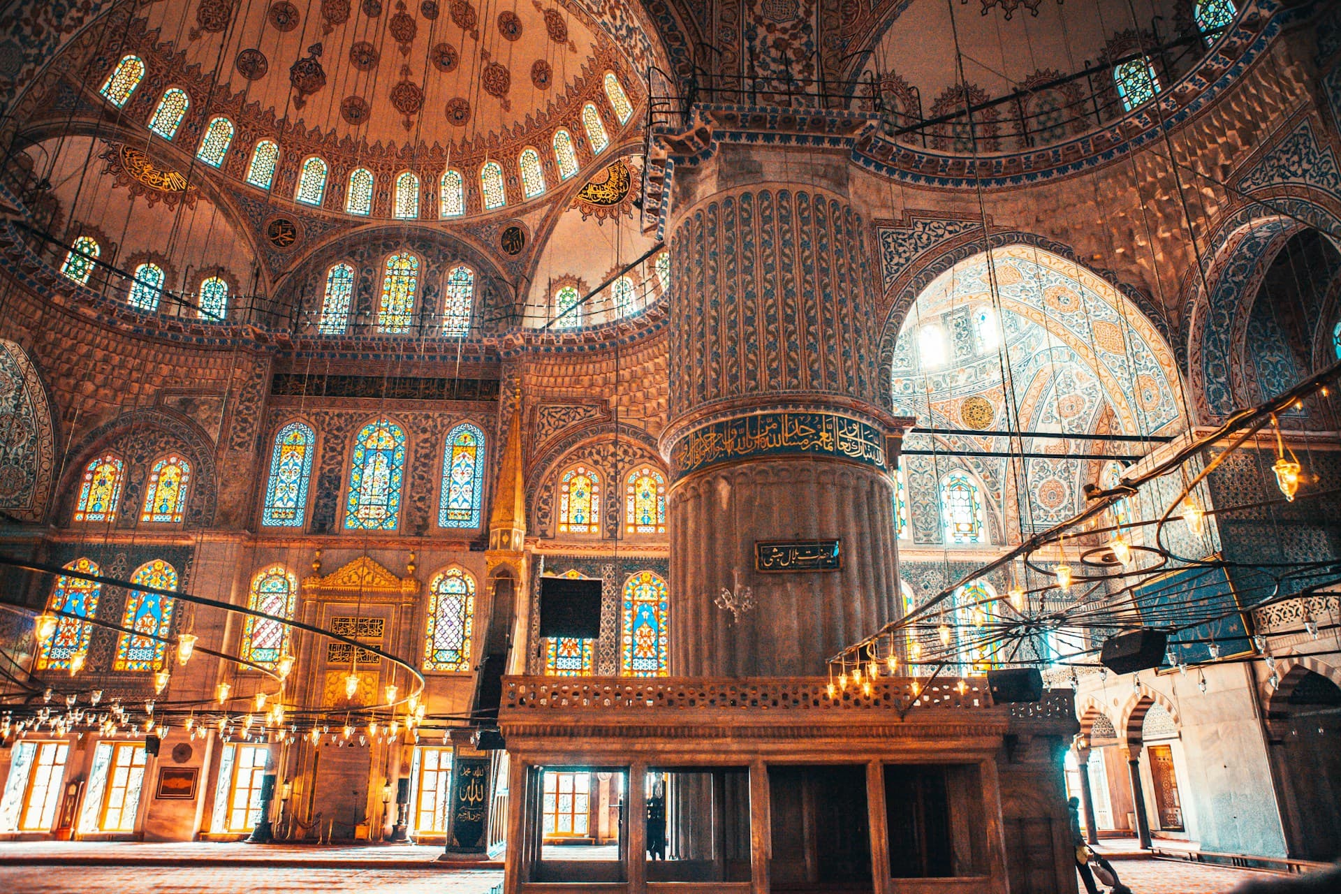 Ornate historic mosque interior with decorated domes, tall central column, stained-glass windows and hanging chandeliers.