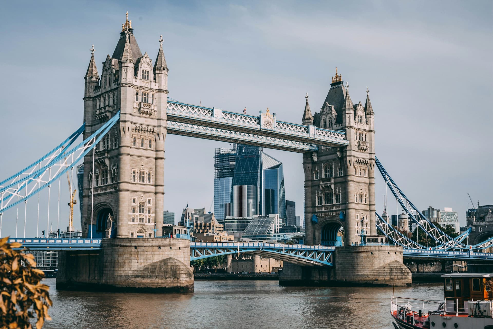 Tower Bridge spanning the River Thames in London with modern skyscrapers in the background under a cloudy sky.