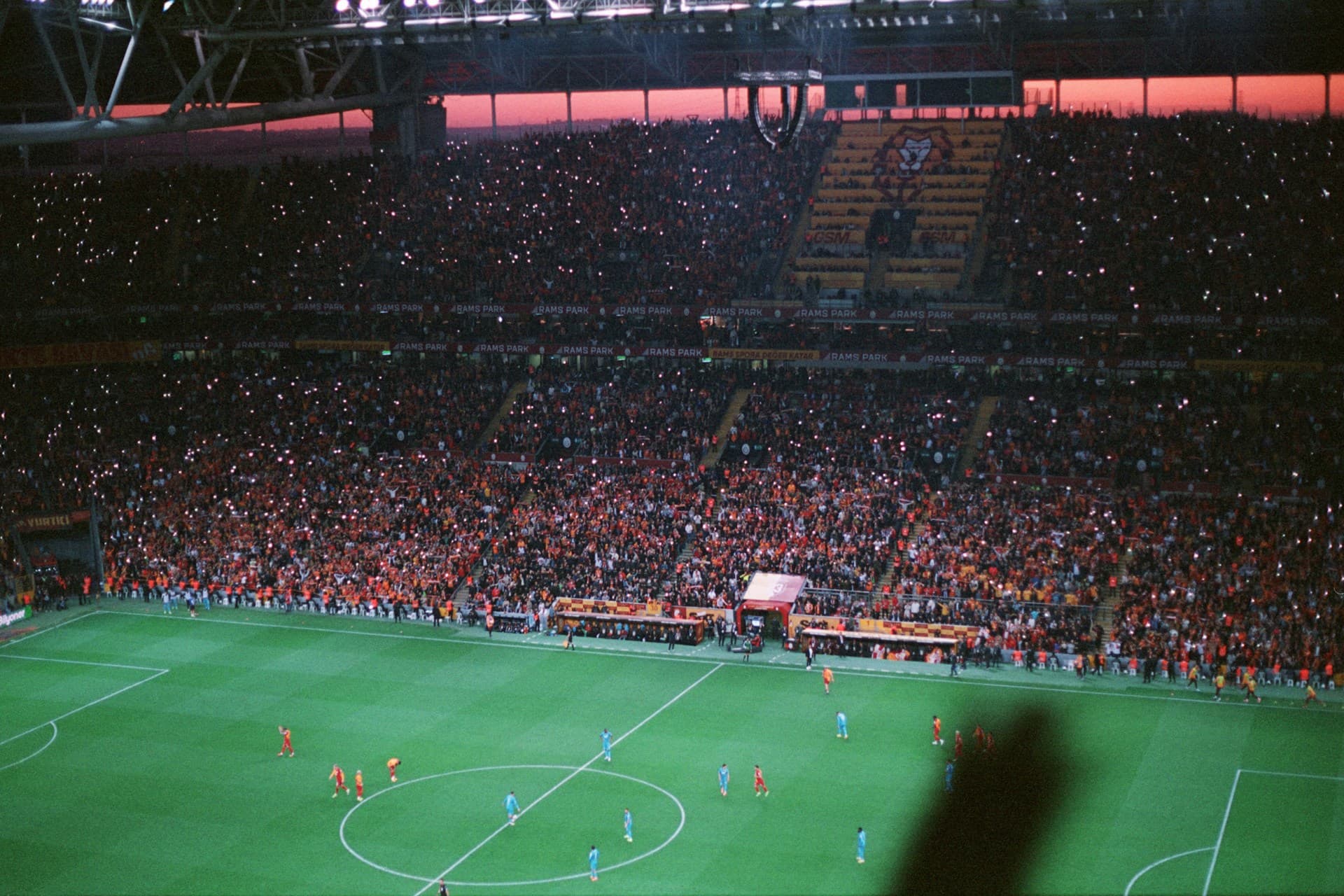 Overhead view of a packed soccer stadium at dusk, fans' lights sparkling as red and blue teams play on the field.