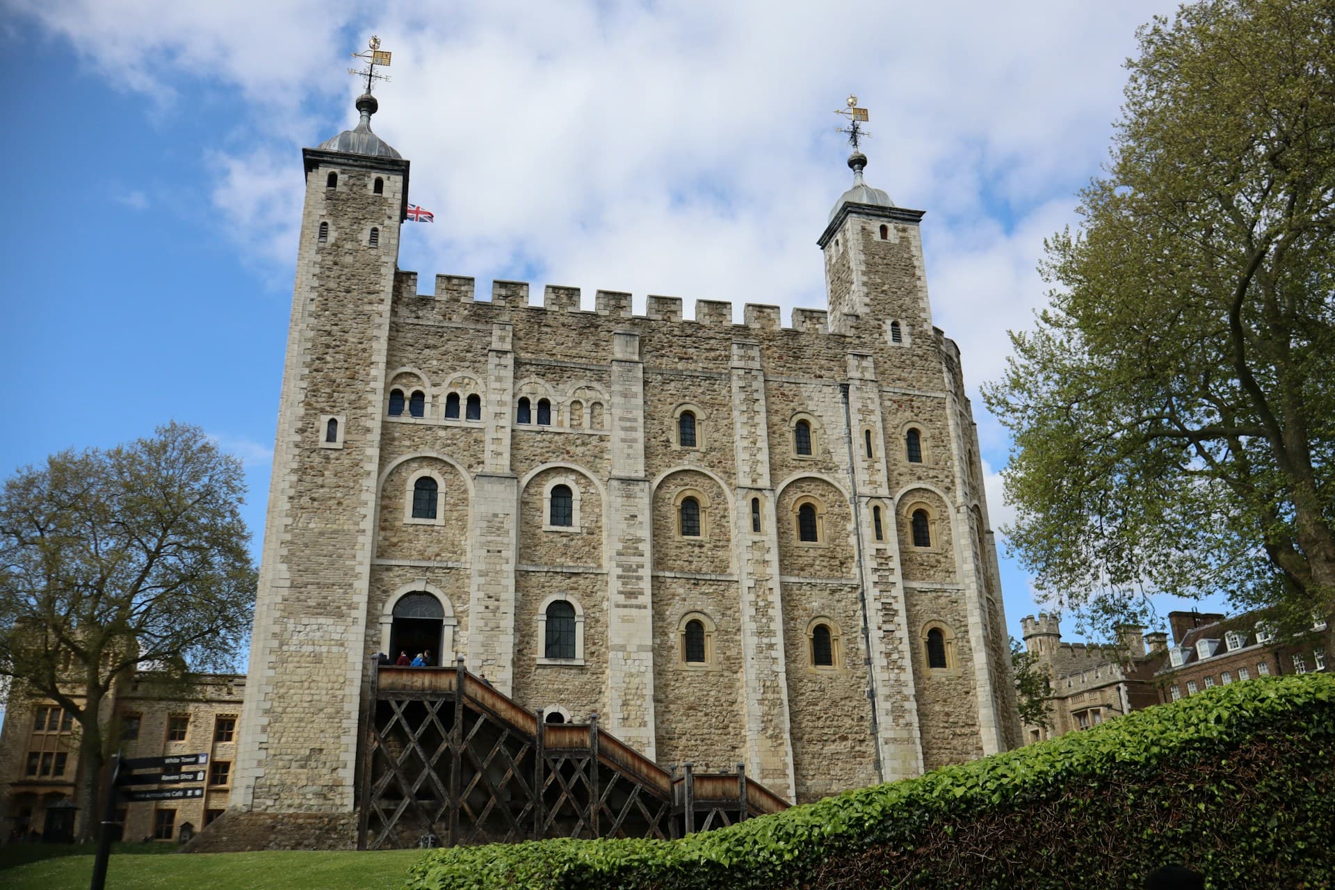 Stone medieval keep with battlements and twin turrets, wooden external staircase, grassy foreground and trees under a partly cloudy sky.