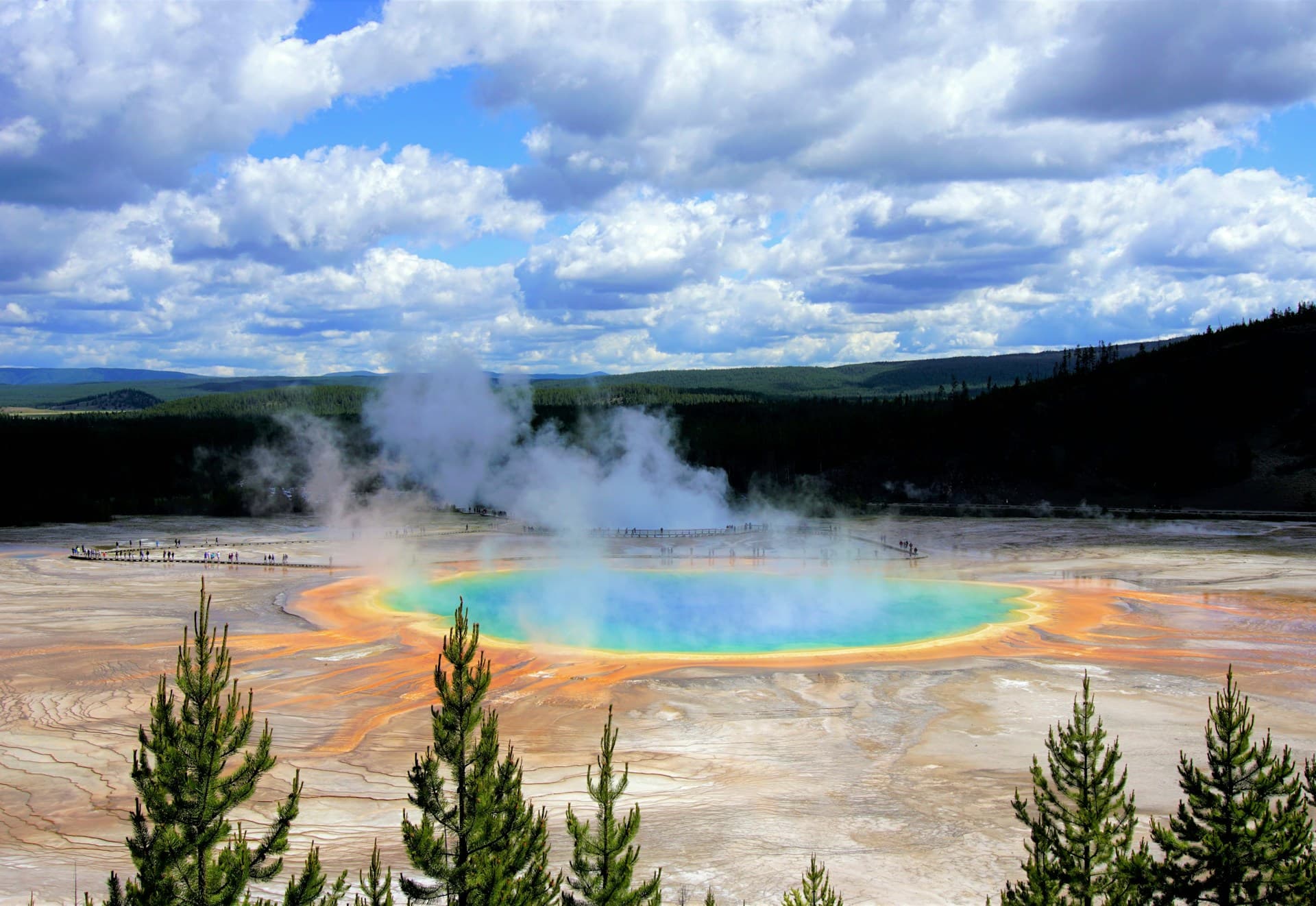 A vibrant geothermal spring with blue, green, and orange hues surrounded by steam, set against a forested landscape and cloudy sky.