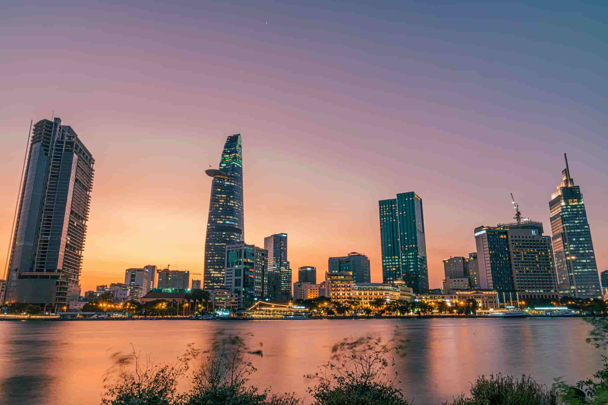 City skyline at dusk with illuminated skyscrapers reflecting in a calm river, against a gradient sky transitioning from orange to purple.