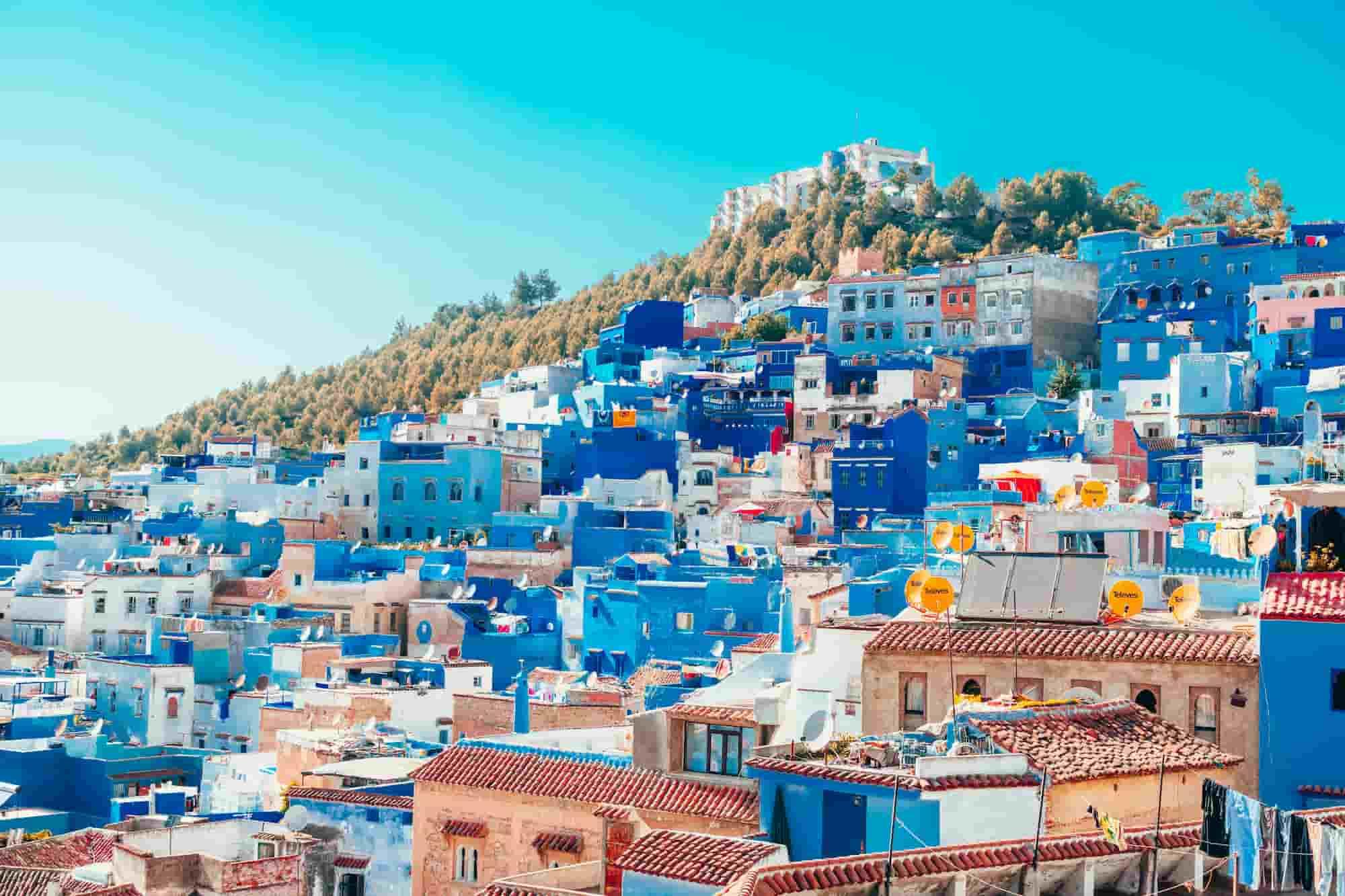 A vibrant hillside town with blue-painted buildings under a clear blue sky, surrounded by lush greenery.