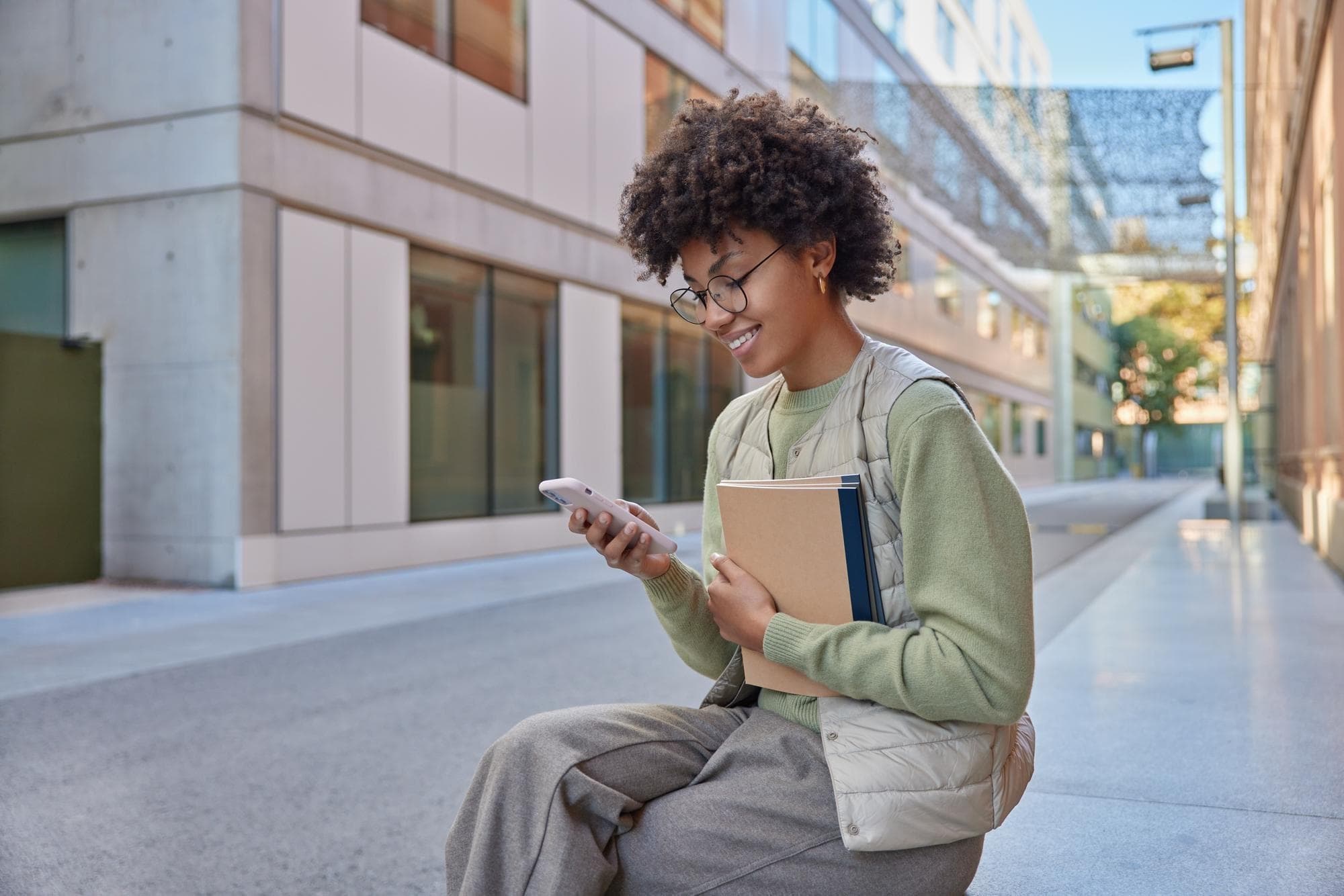 A person with curly hair and glasses smiles at their phone while holding books, seated in an outdoor urban setting.