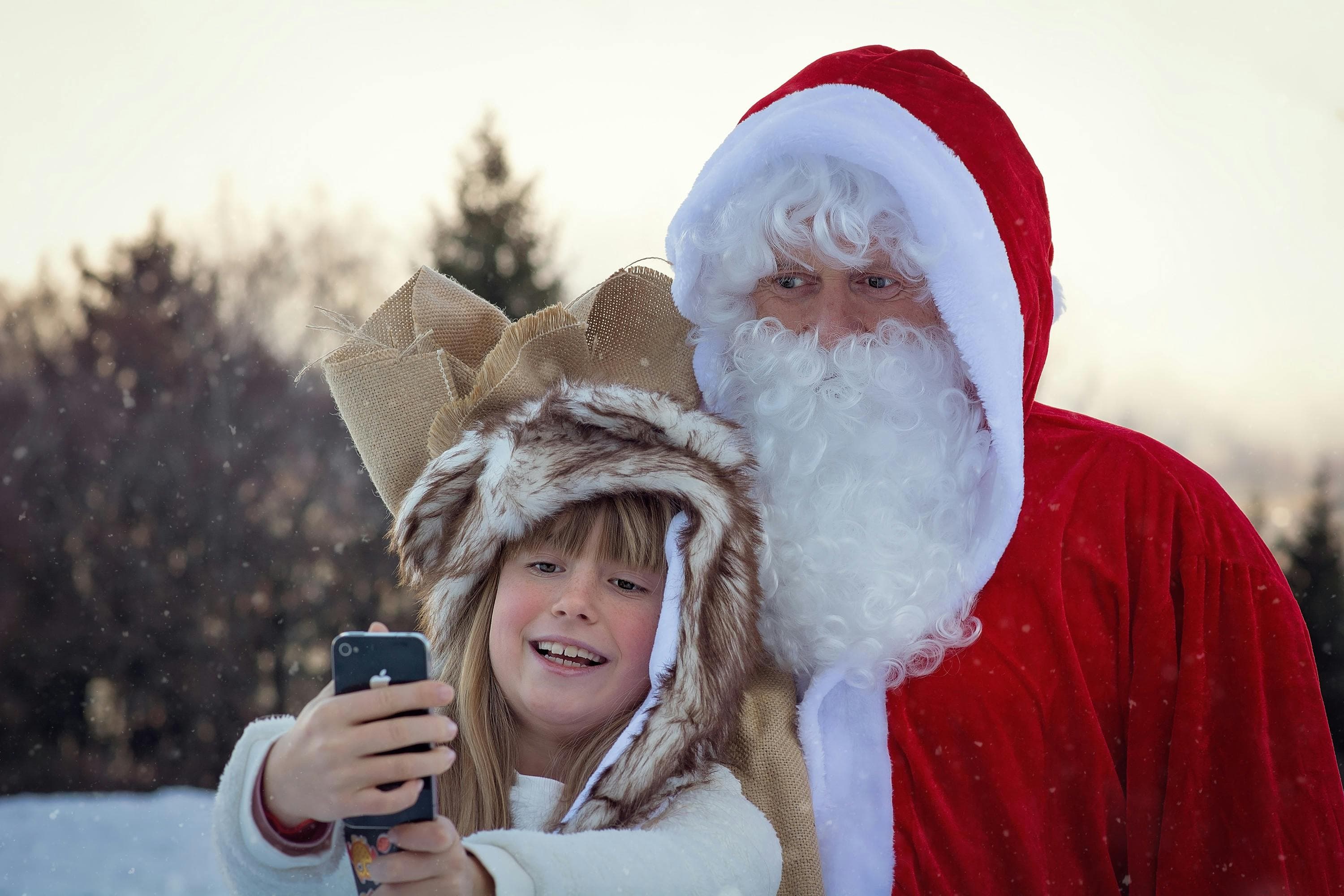 A child in a furry hat takes a selfie with Santa Claus outdoors in a snowy setting, both smiling and enjoying the moment.