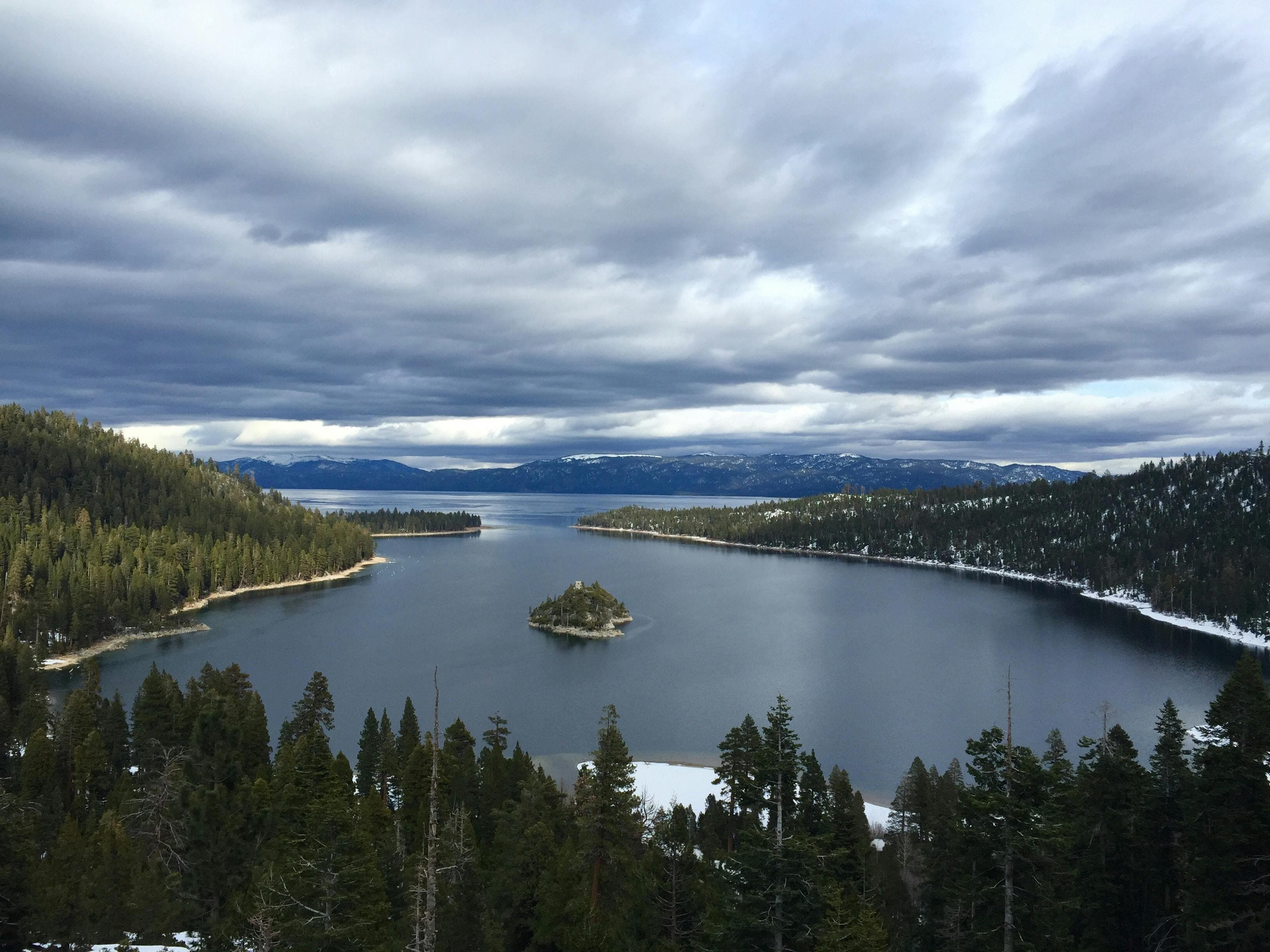 A serene lake surrounded by dense forests and snow-capped mountains under a cloudy sky, with a small island in the center.