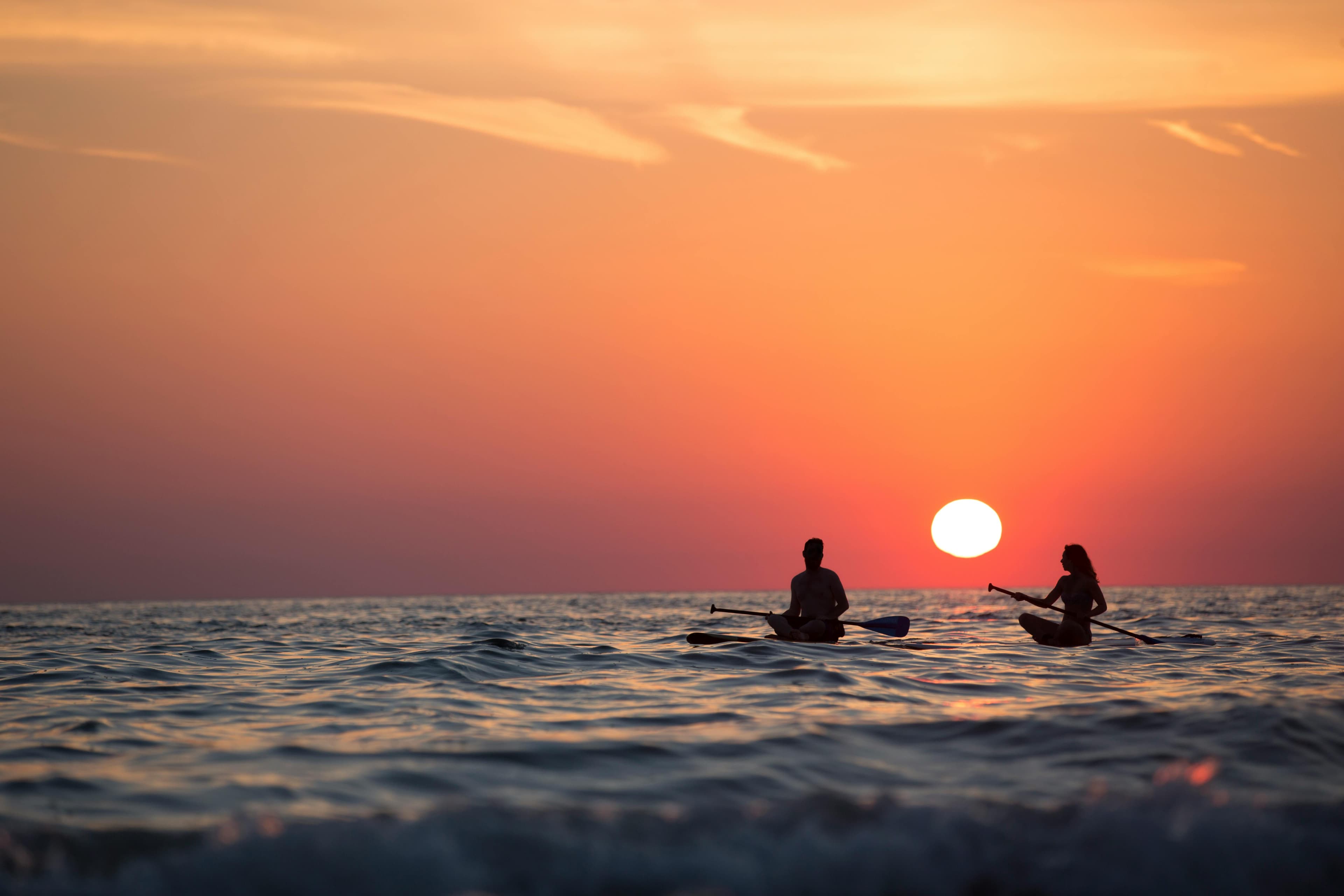 Two people kayaking on the ocean at sunset, with an orange sky and the sun near the horizon.