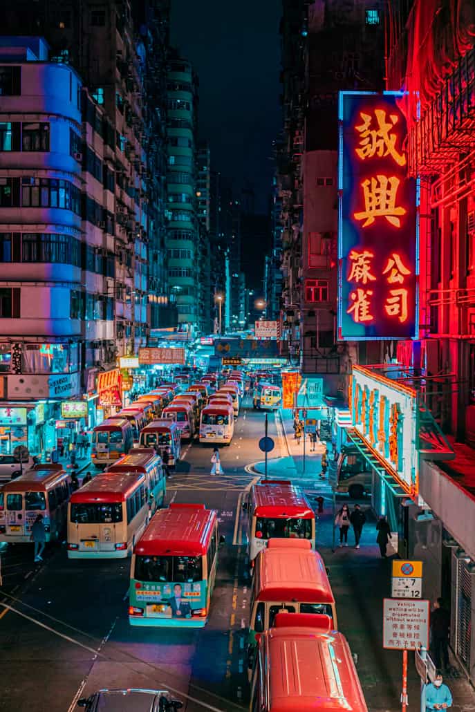 Bustling city street at night with vibrant neon signs and red minibuses, flanked by tall buildings under a dark sky.