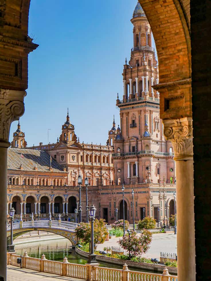 Ornate building and bell tower framed by an arch, with a bridge and river in the foreground under a clear blue sky.