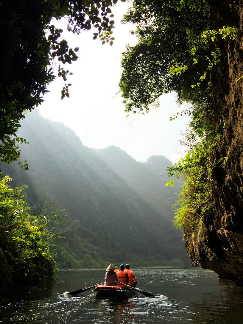 Two people rowing a small boat through a tranquil river gorge surrounded by lush green cliffs under soft sunlight filtering through the foliage.